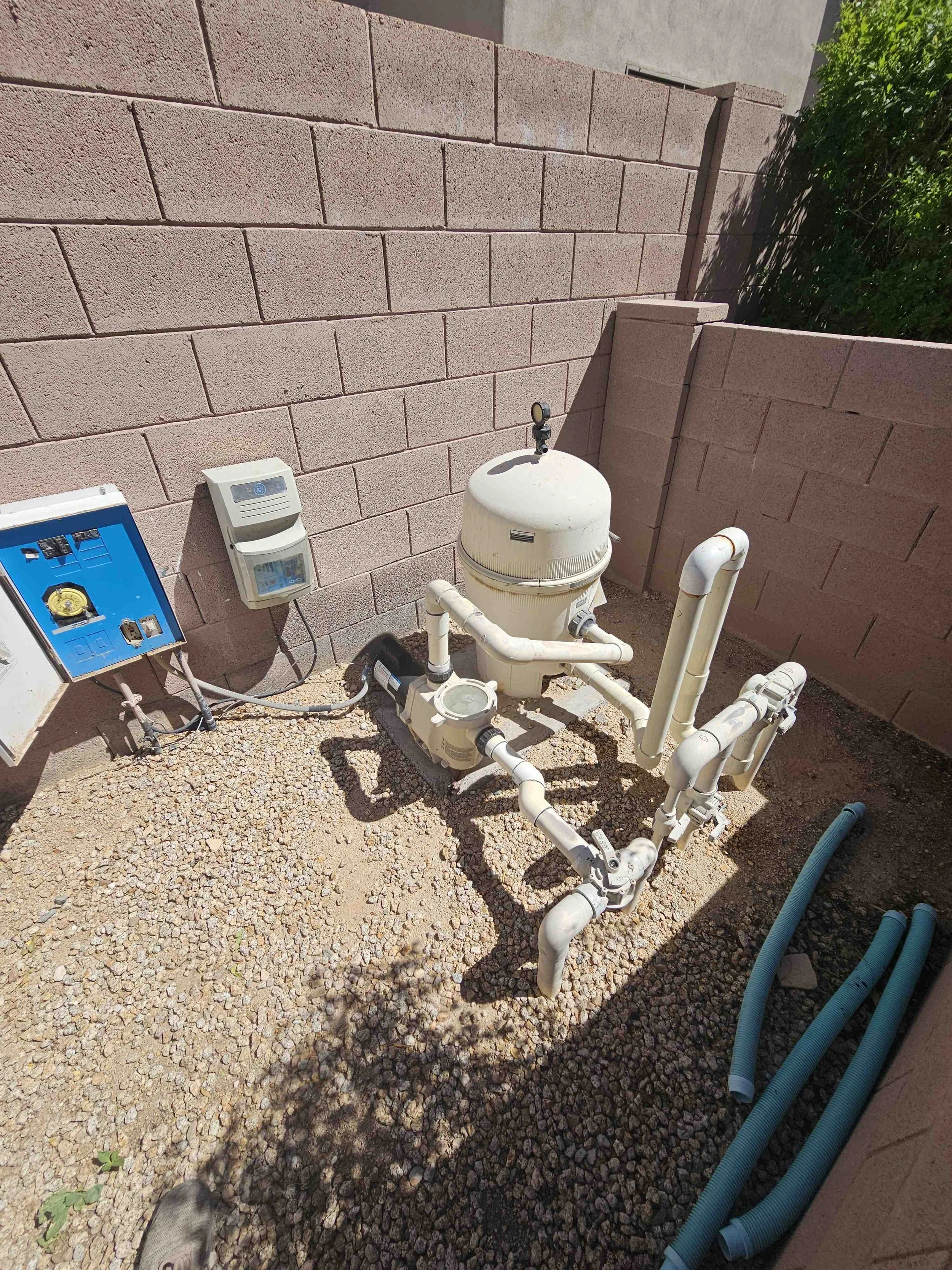 Close-up view of a small outdoor plumbing setup with white pipes, a filter, and an electrical control box against a pink brick wall, with gravel ground and some green foliage on the side.