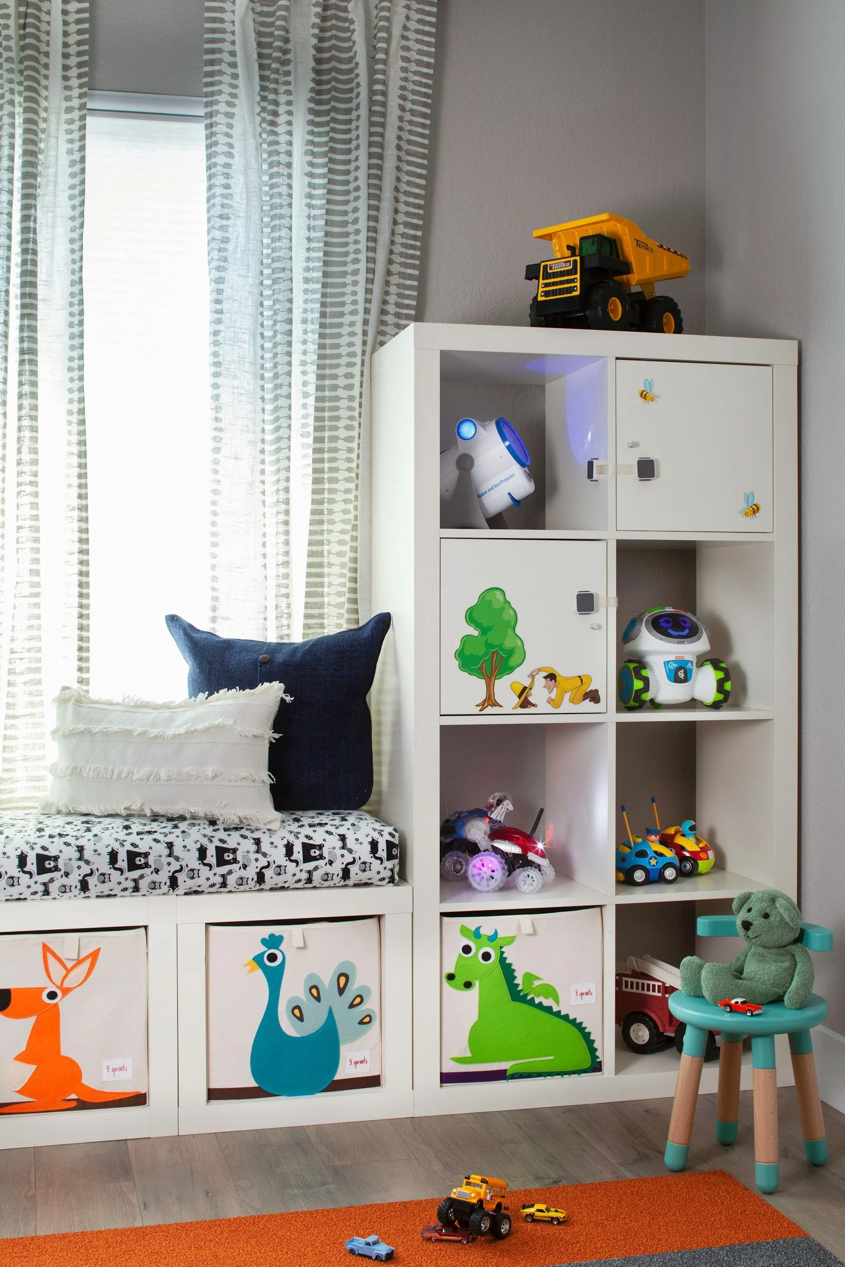 Child's playroom with a white storage unit decorated with animal illustrations, a window with curtains, Toy vehicles, stuffed animals, and a chair with a green teddy bear, toys on the orange rug.
