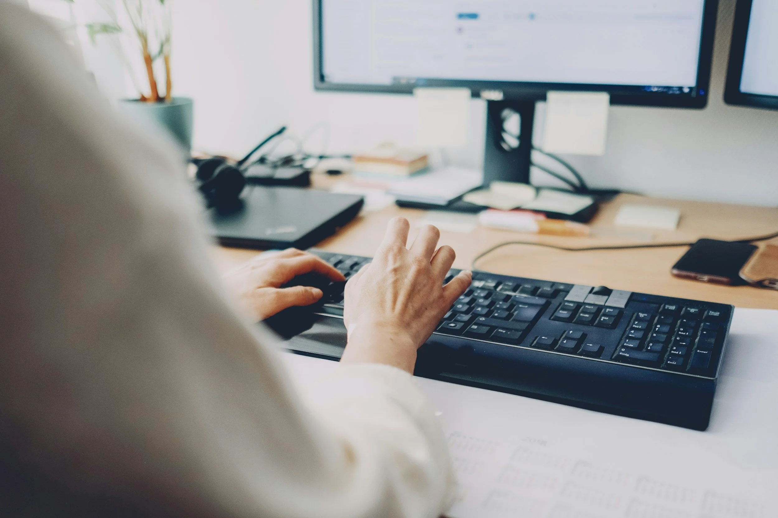 A person typing on a black computer keyboard in an office space, with a large monitor displaying a content management system or social media platform in the background, along with a laptop, some sticky notes, and a smartphone on the desk.