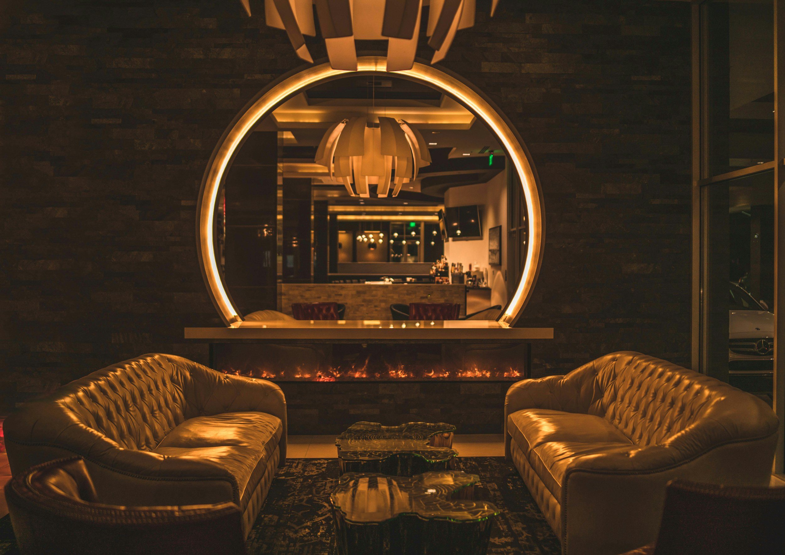 A luxurious hotel lobby featuring leather couches, a large illuminated circular mirror, modern chandeliers, and a dark textured wall.