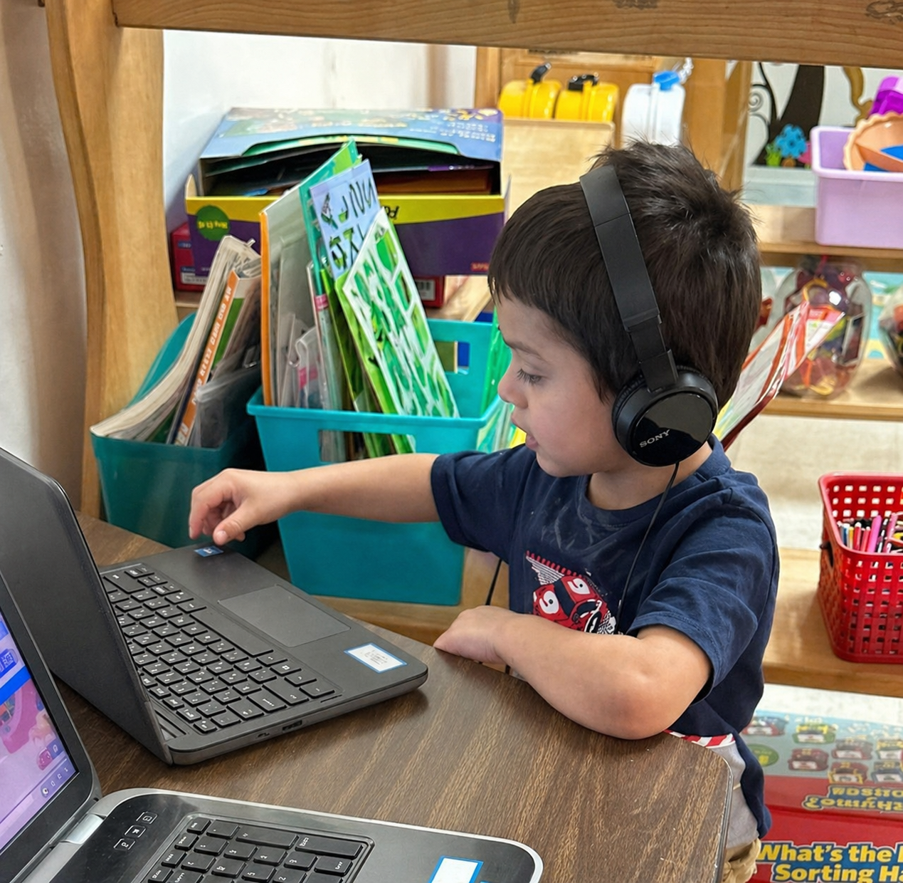 A young boy with dark hair wearing a blue t-shirt with a cartoon character, sitting at a wooden table with two laptops, wearing black headphones, in a classroom setting with colorful books and supplies.
