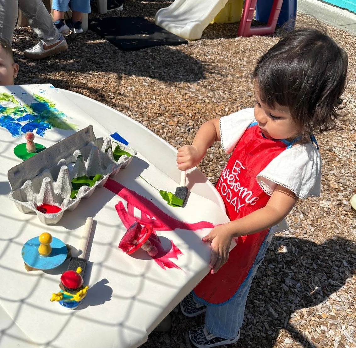 Child painting with red and green paint at a white table outdoors on a playground.