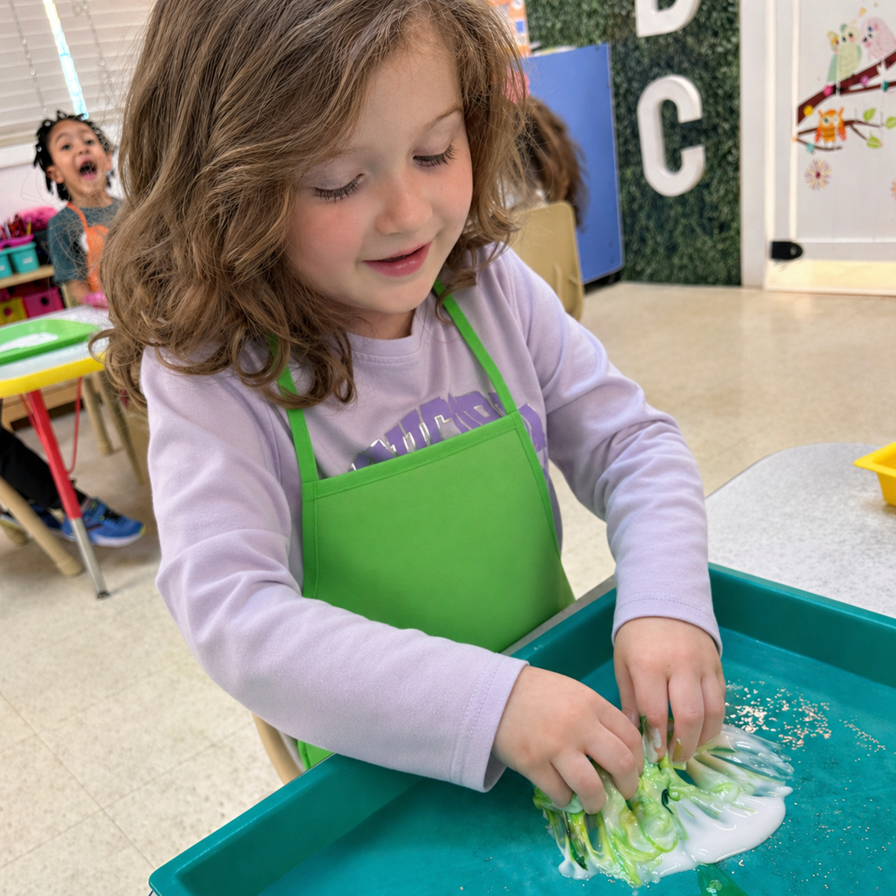 A young girl with curly hair wearing a purple shirt and green apron playing with colorful slime at a table in a classroom.
