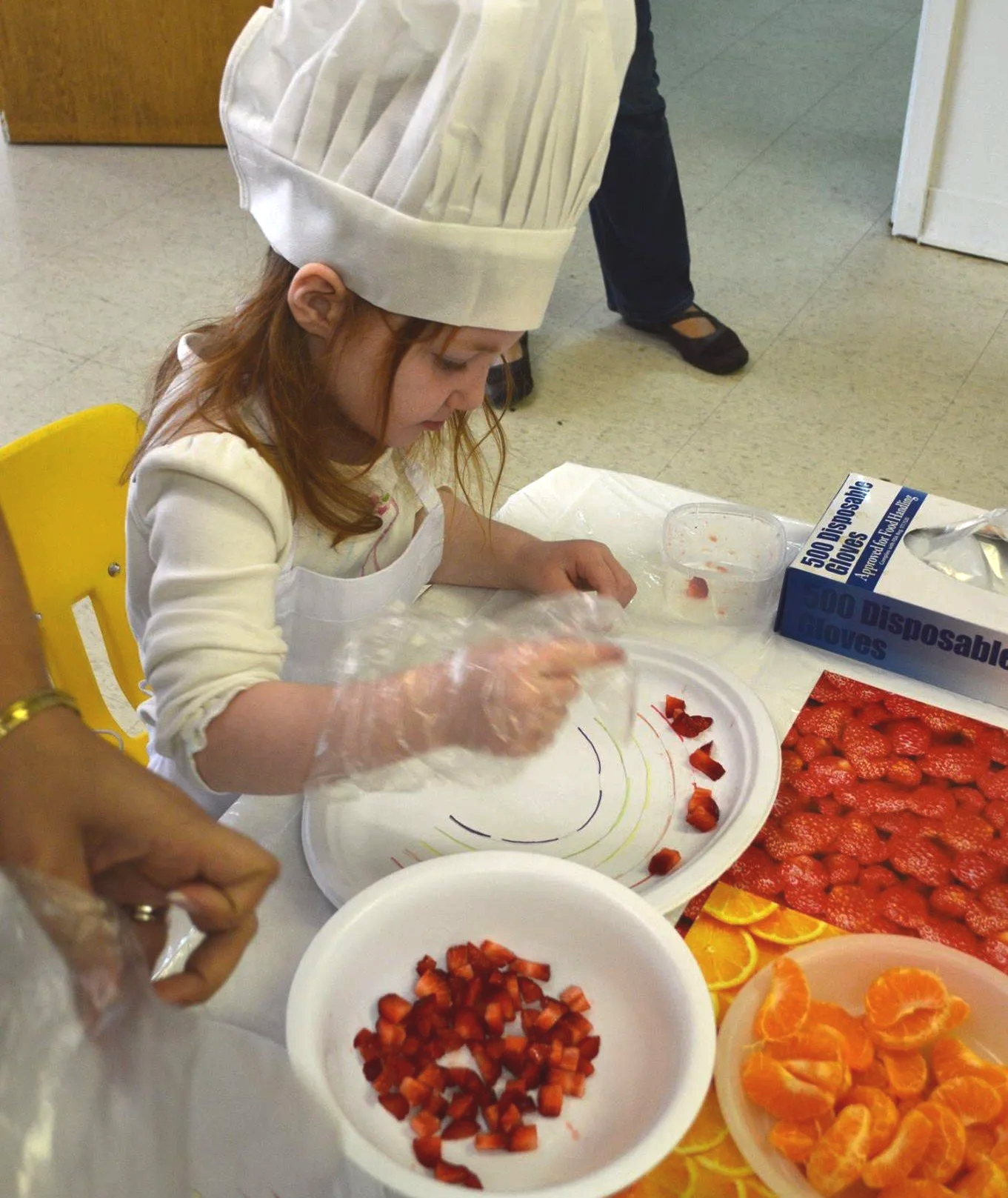 Young girl wearing a chef's hat and apron, preparing strawberries and oranges at a table with various fruit trays, including sliced strawberries and orange segments.