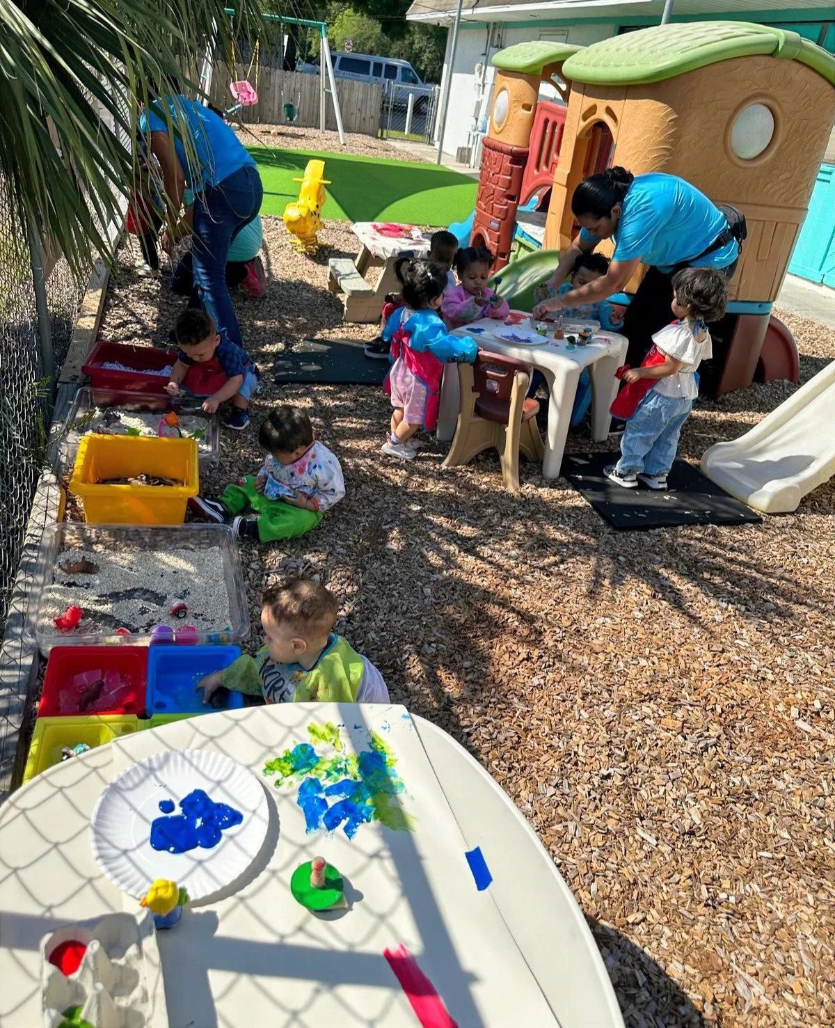 Children playing outside in a backyard with toys and a playhouse, supervised by adults, some engaging in painting and others playing in sensory bins.