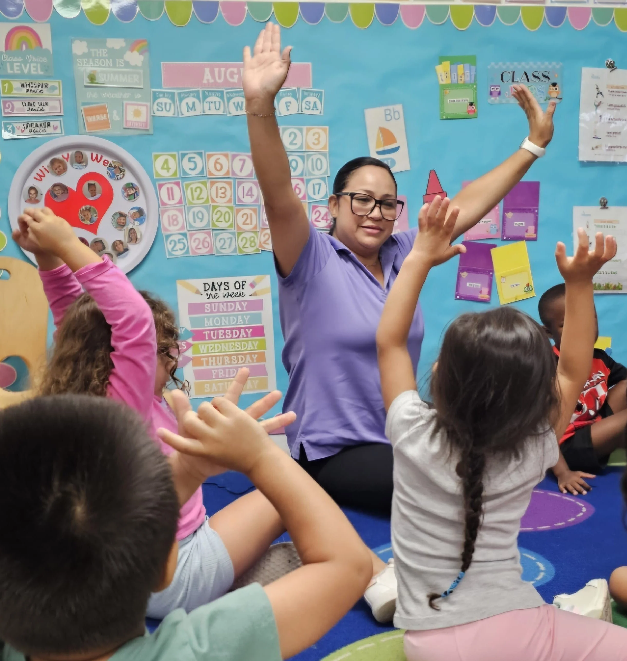A group of young children, some with their hands raised, participate in a classroom activity led by a woman wearing glasses and a purple shirt. The classroom background features colorful educational posters, including a calendar, days of the week, and other learning materials.