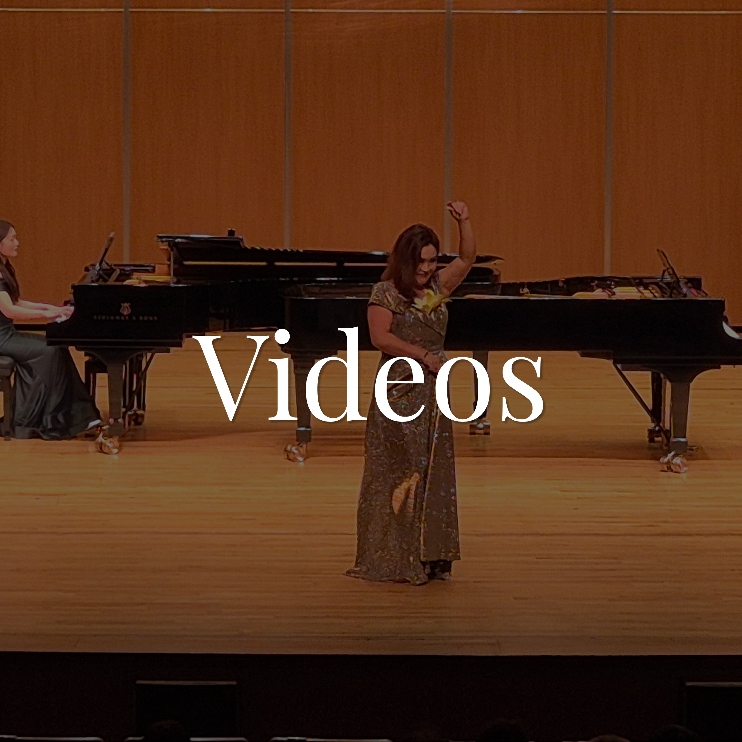 Woman with long hair and glasses standing at a concert or recital hall, raising her arm in celebration or applause, with a grand piano and other musicians visible in the background.