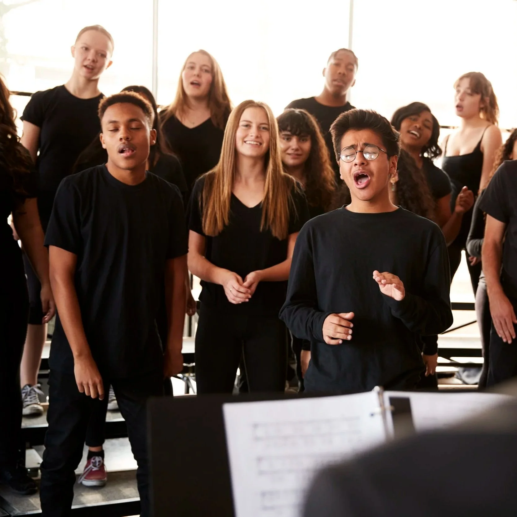 A group of people singing in a choir, with a focus on two young women in the front and others in the background, all wearing black clothing, inside a bright room.