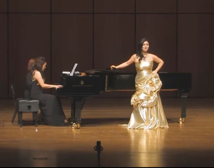 A woman playing the piano and another woman standing next to a grand piano on a stage in a concert hall.