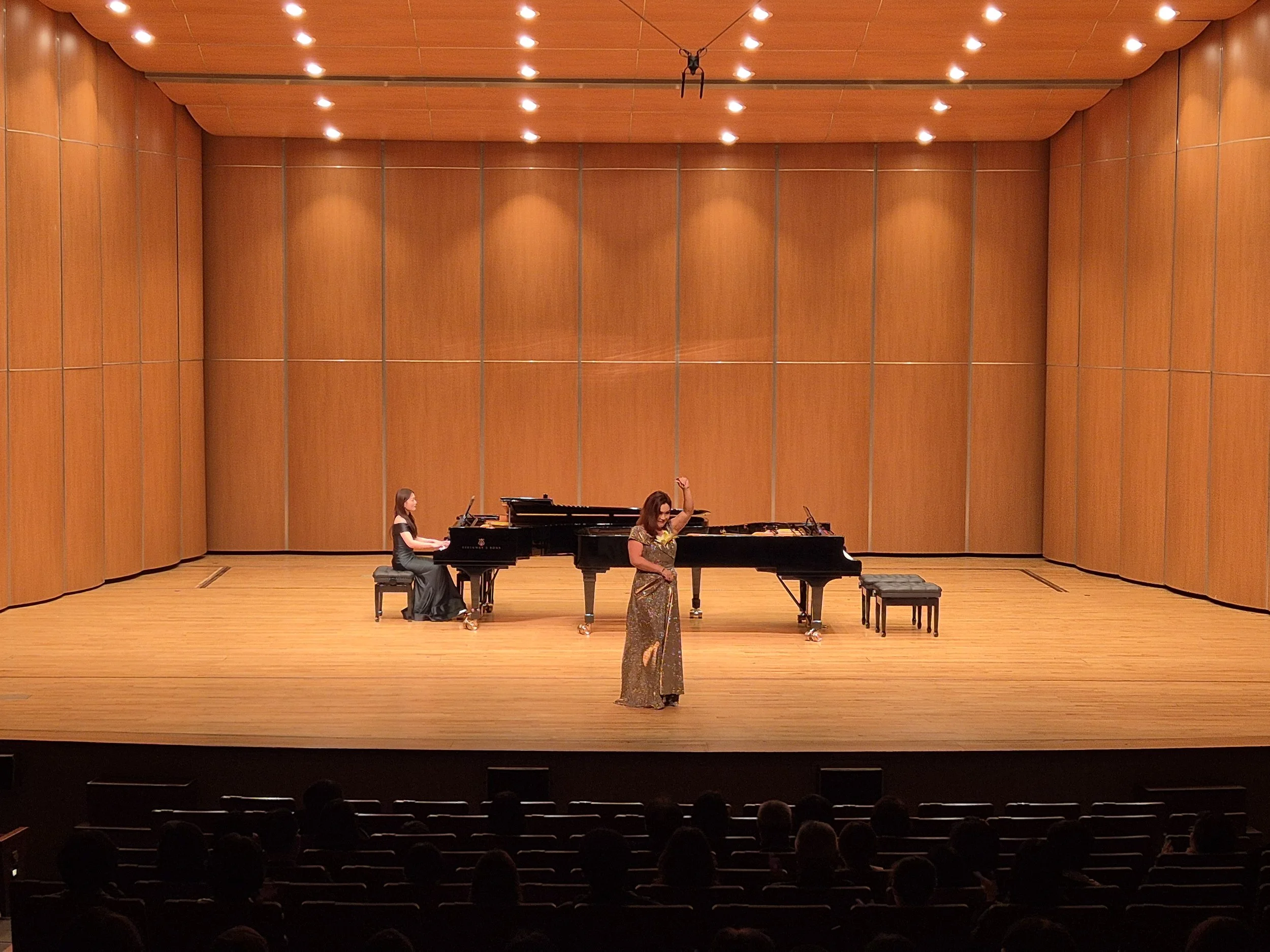 Two women performing on stage with concert grand pianos in a concert hall with wooden wall panels. One woman is sitting at a piano, and the other is standing and gesturing with her hand.