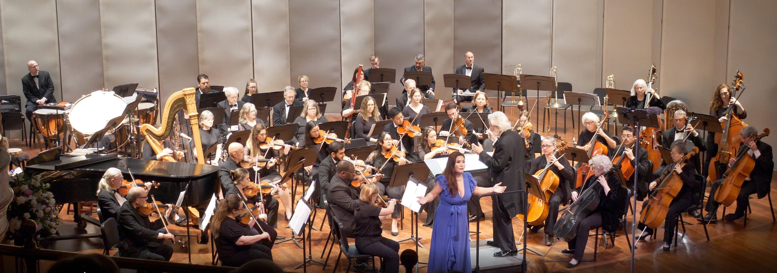 An orchestra performing on stage with musicians playing various string, wind, and percussion instruments, led by a conductor, in a concert hall.