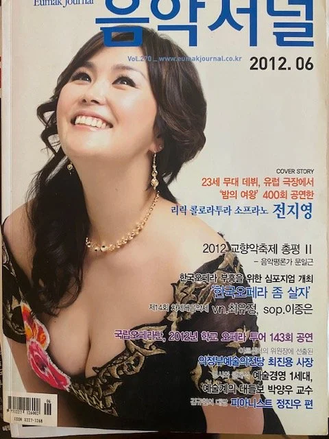 A magazine cover featuring a smiling woman with dark, curly hair, wearing a black dress with colorful floral accents and jewelry, including earrings and a necklace, with Korean text and the date June 2012.