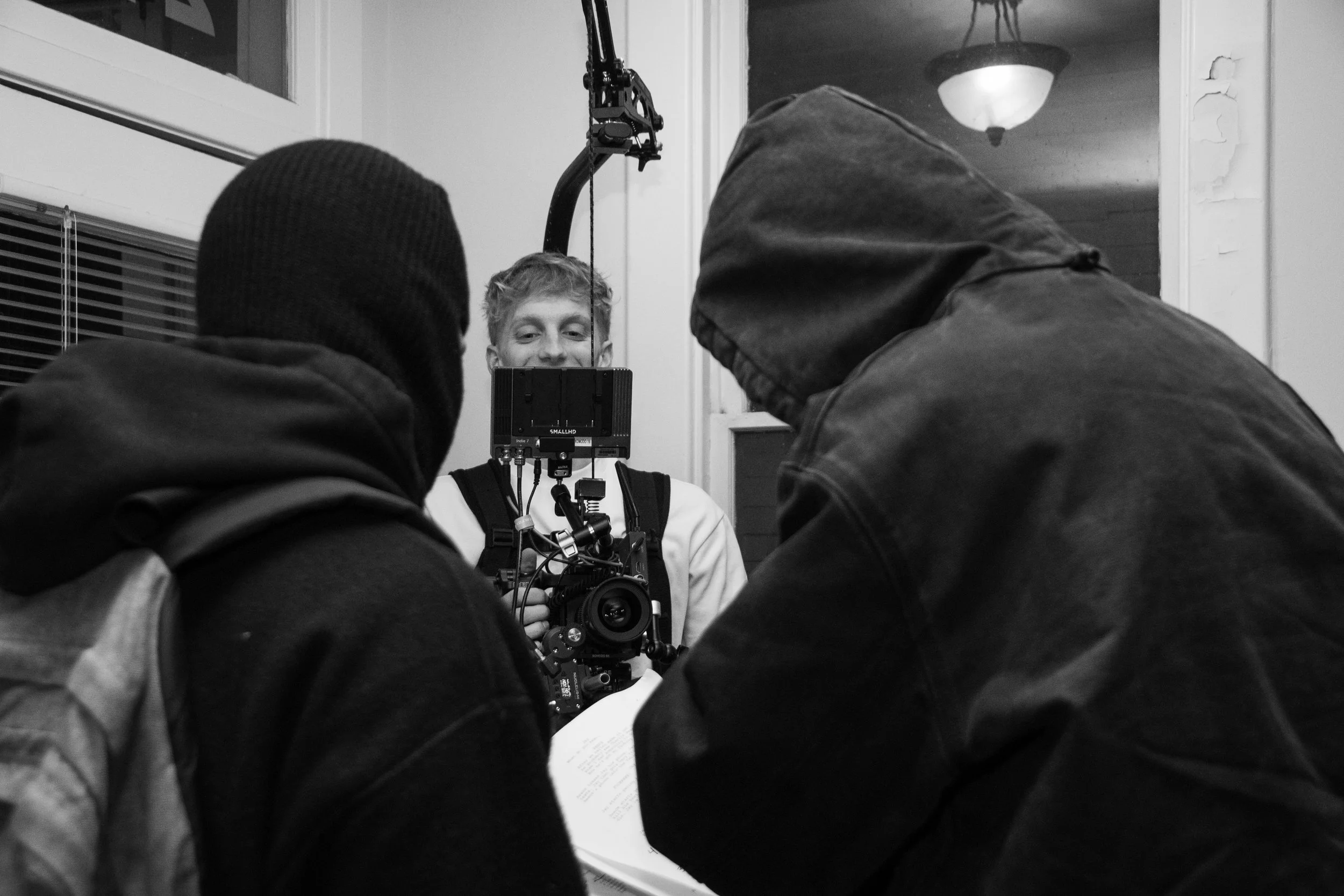 Three people involved in a film shoot, with one holding a camera and the other two wearing hooded jackets, one of them reading from a script, inside a room with a window and a ceiling light.