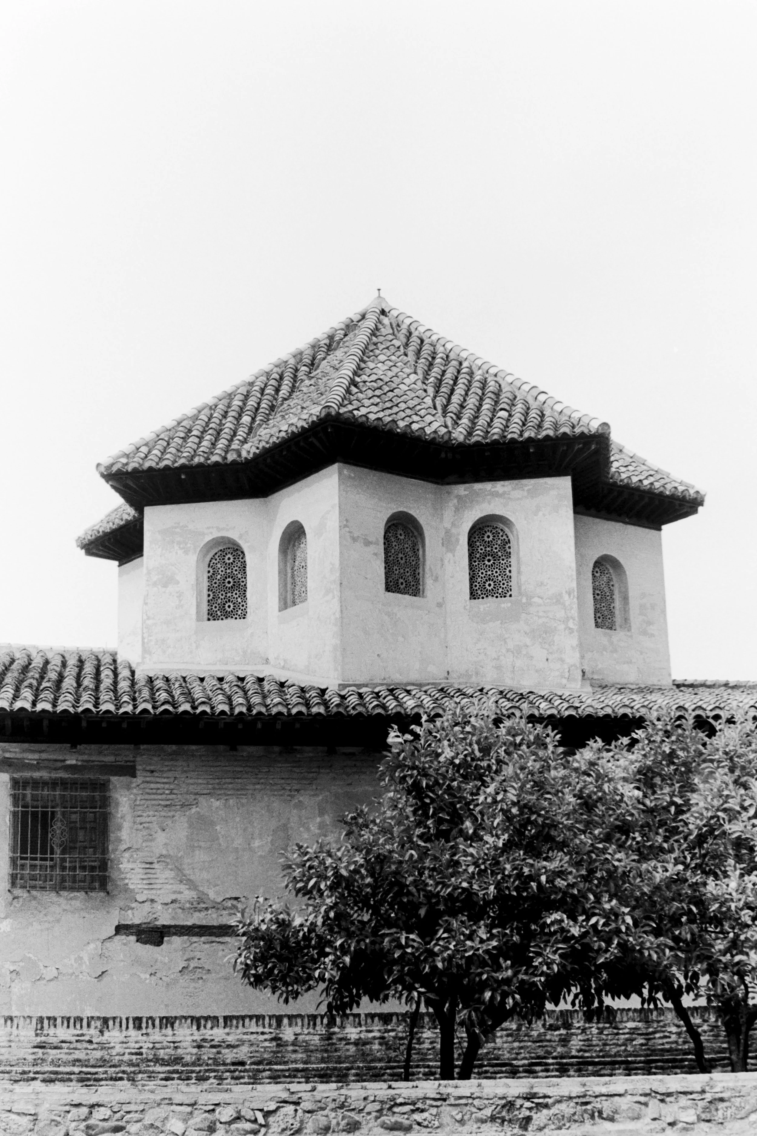 A black and white photograph of a building with a tiled, pyramid-shaped roof and arched windows, partially obscured by a tree at the front.