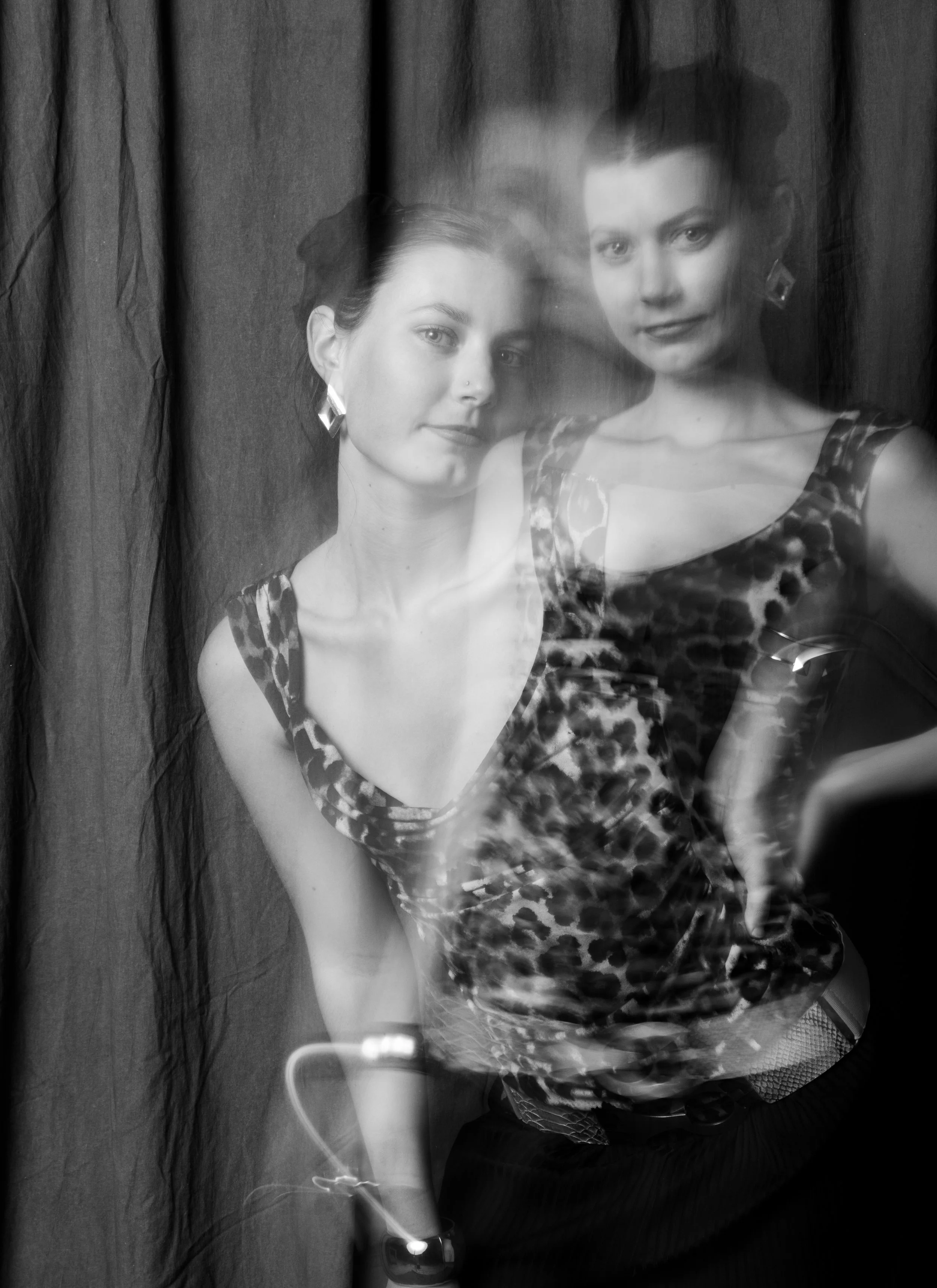 Black and white photo of a woman with dark hair and earrings, wearing a leopard-print top, posing with a hand on her hip, seen through a double exposure or reflection.