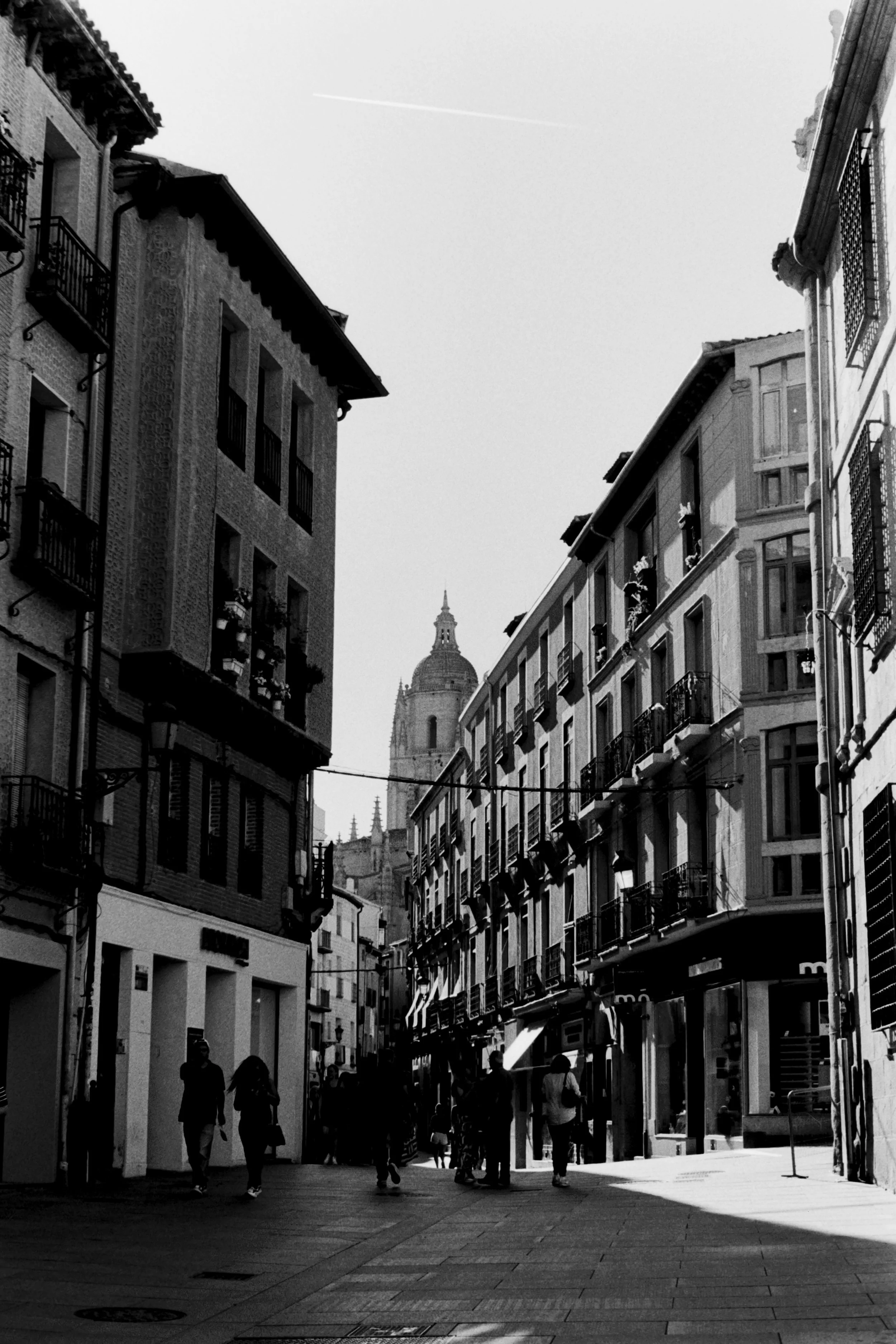Black and white photo of a narrow city street with tall buildings on both sides. People are walking along the sidewalk, and there is a large domed building with a spire visible in the background.