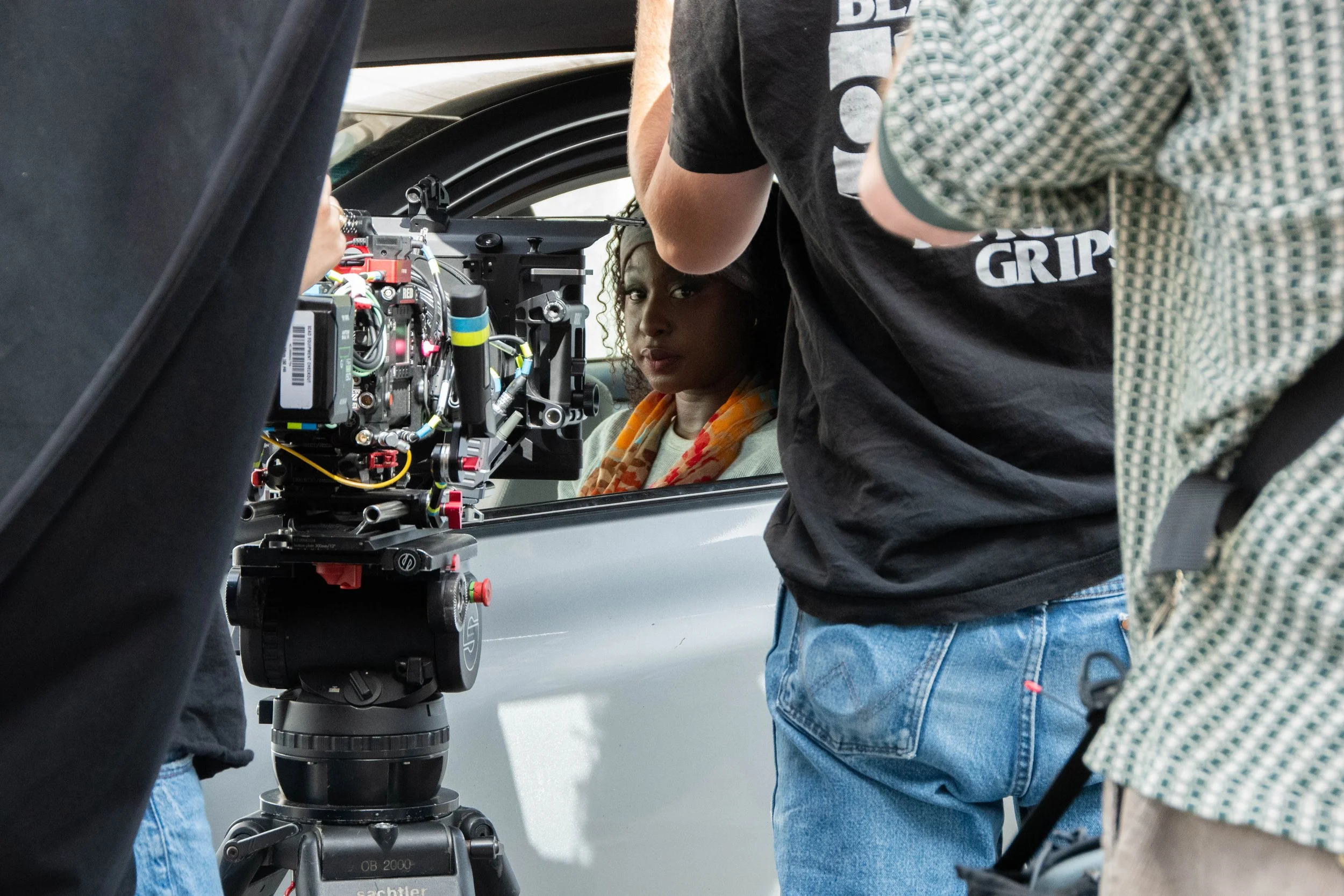 A woman is sitting inside a car, looking out through the window while a film crew films her with professional camera equipment.