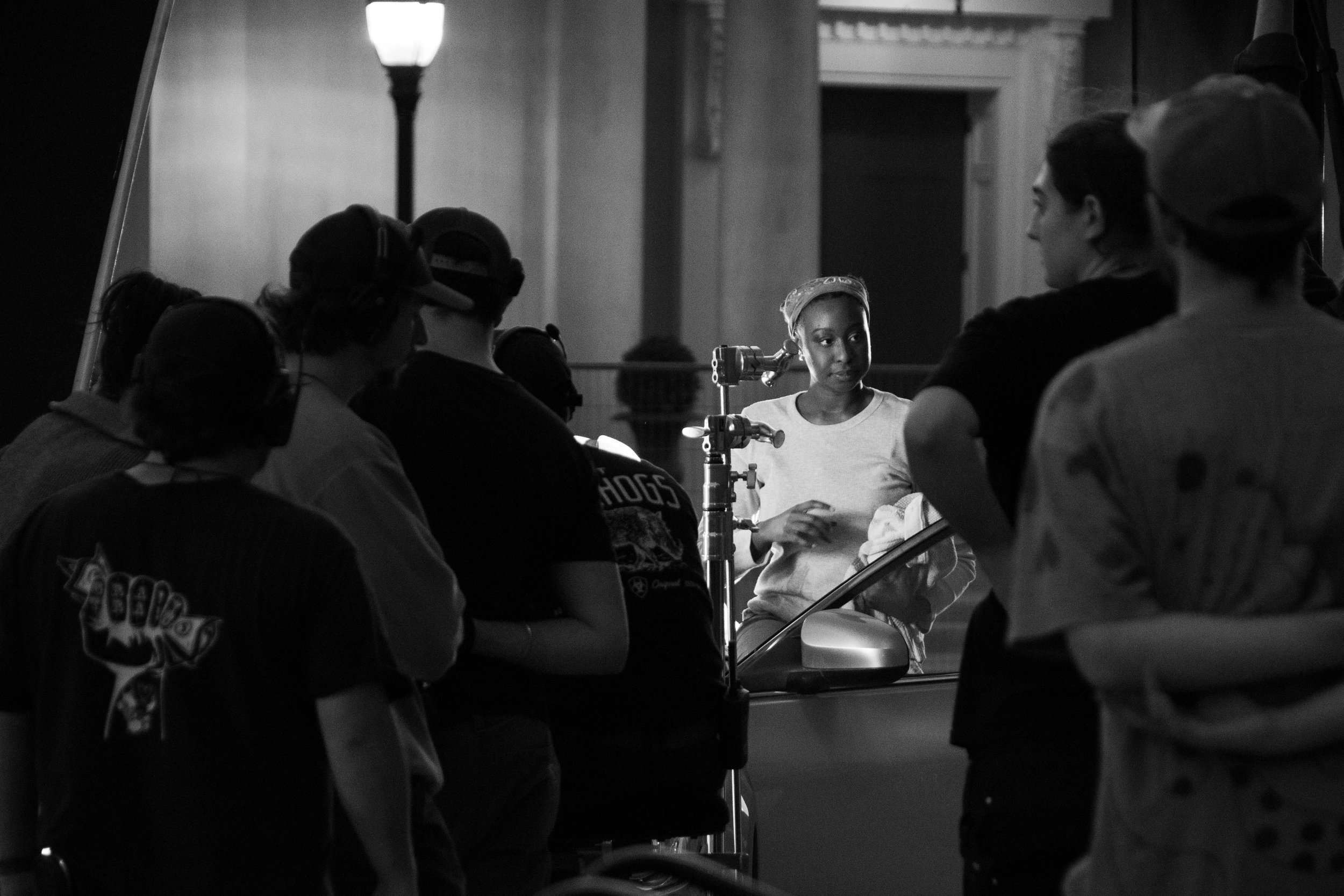 A group of people filming or taking photographs of a young woman with a headscarf, who is standing alongside a car and looking at someone or something outside the frame, illuminated by a streetlamp in an indoor setting.