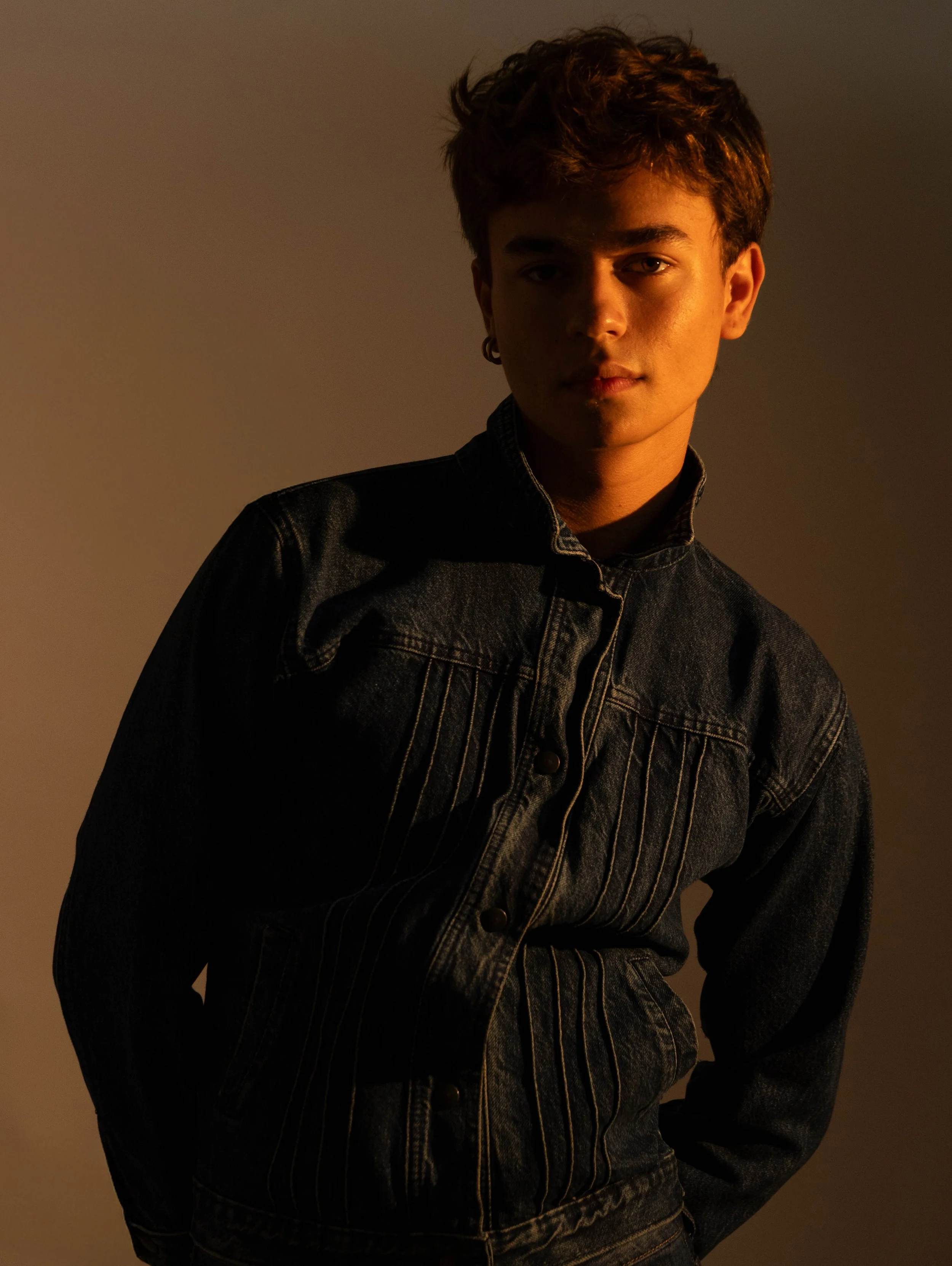 A person with short, wavy hair wearing a dark denim jacket, standing against a neutral background with lighting creating shadowed and illuminated areas on the face and jacket.