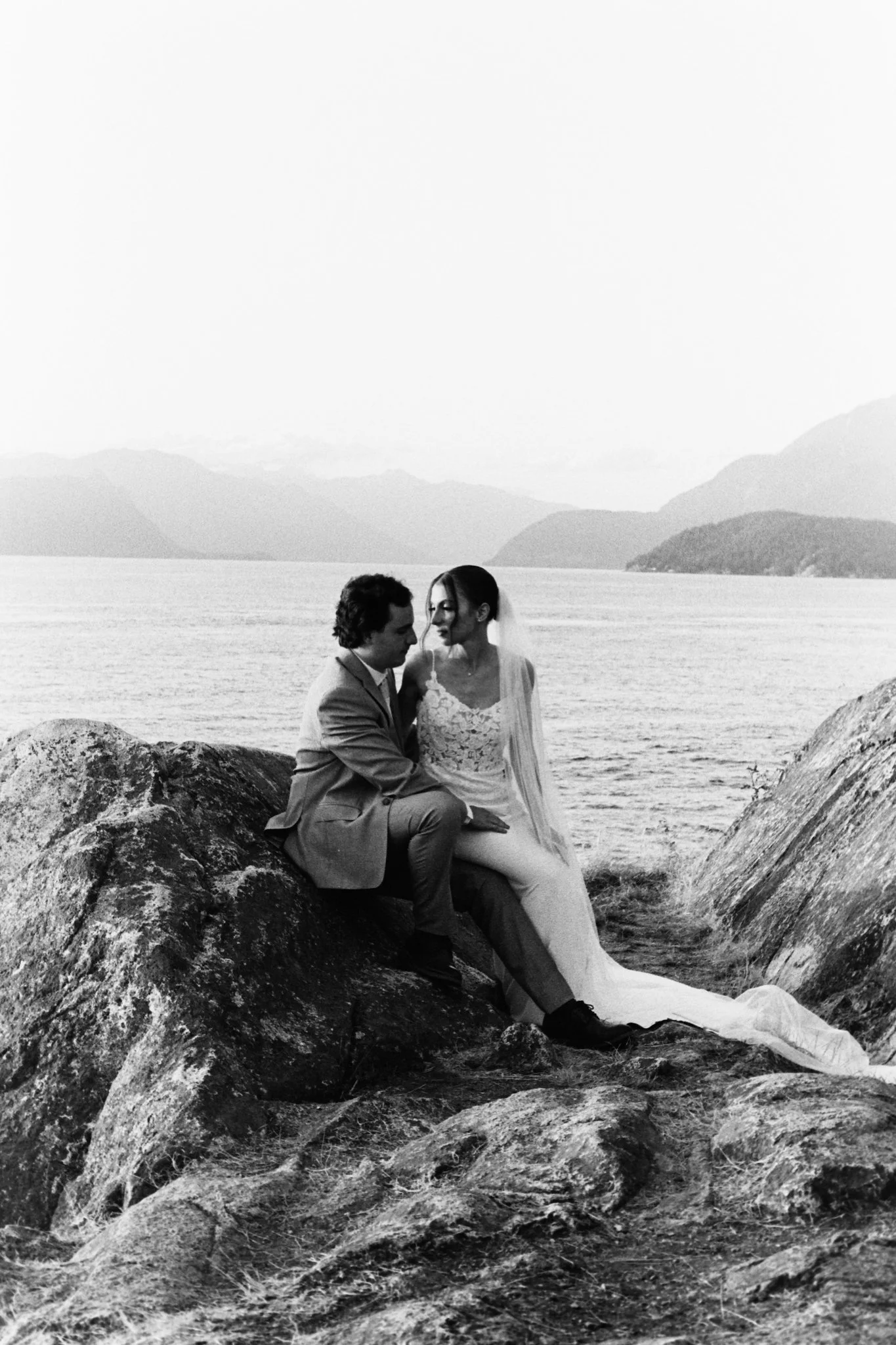 A black and white photo of a bride and groom sitting on rocks near a body of water with mountains in the background. They are looking at each other, dressed in wedding attire.