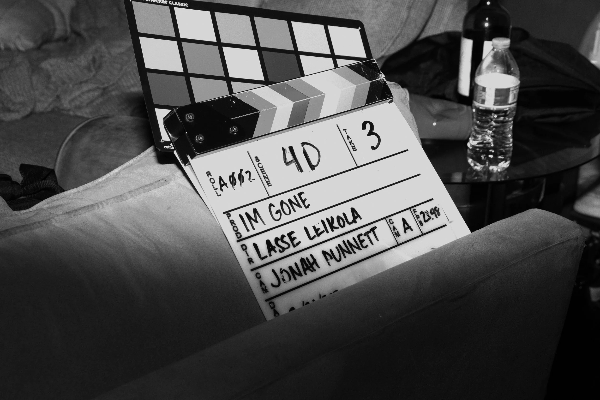 Clapperboard on a sofa with a script written on it, next to a plastic water bottle and a glass of water on a glass table in a living room.