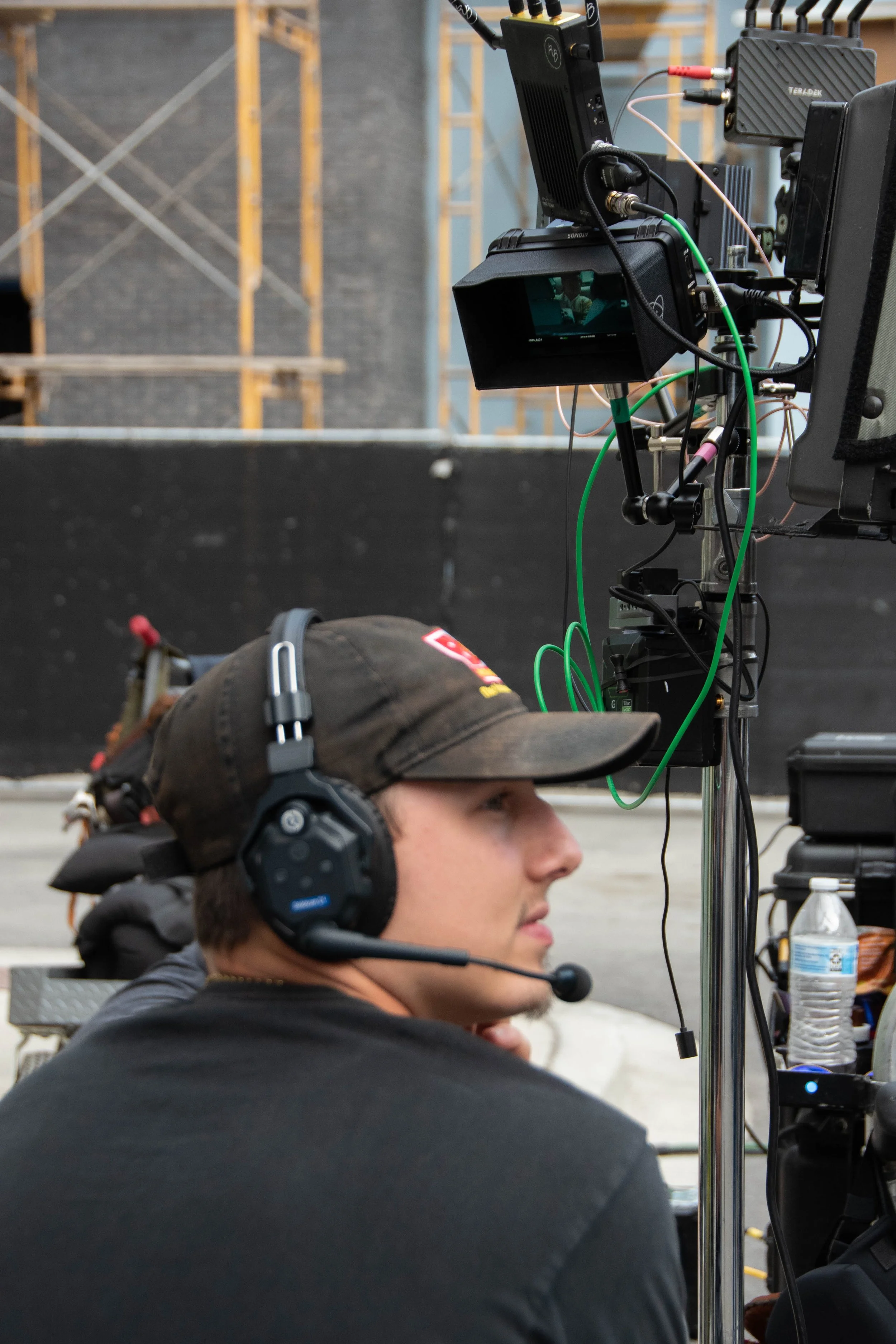 A man wearing a headset and baseball cap sitting in front of a filming camera setup on a film set.