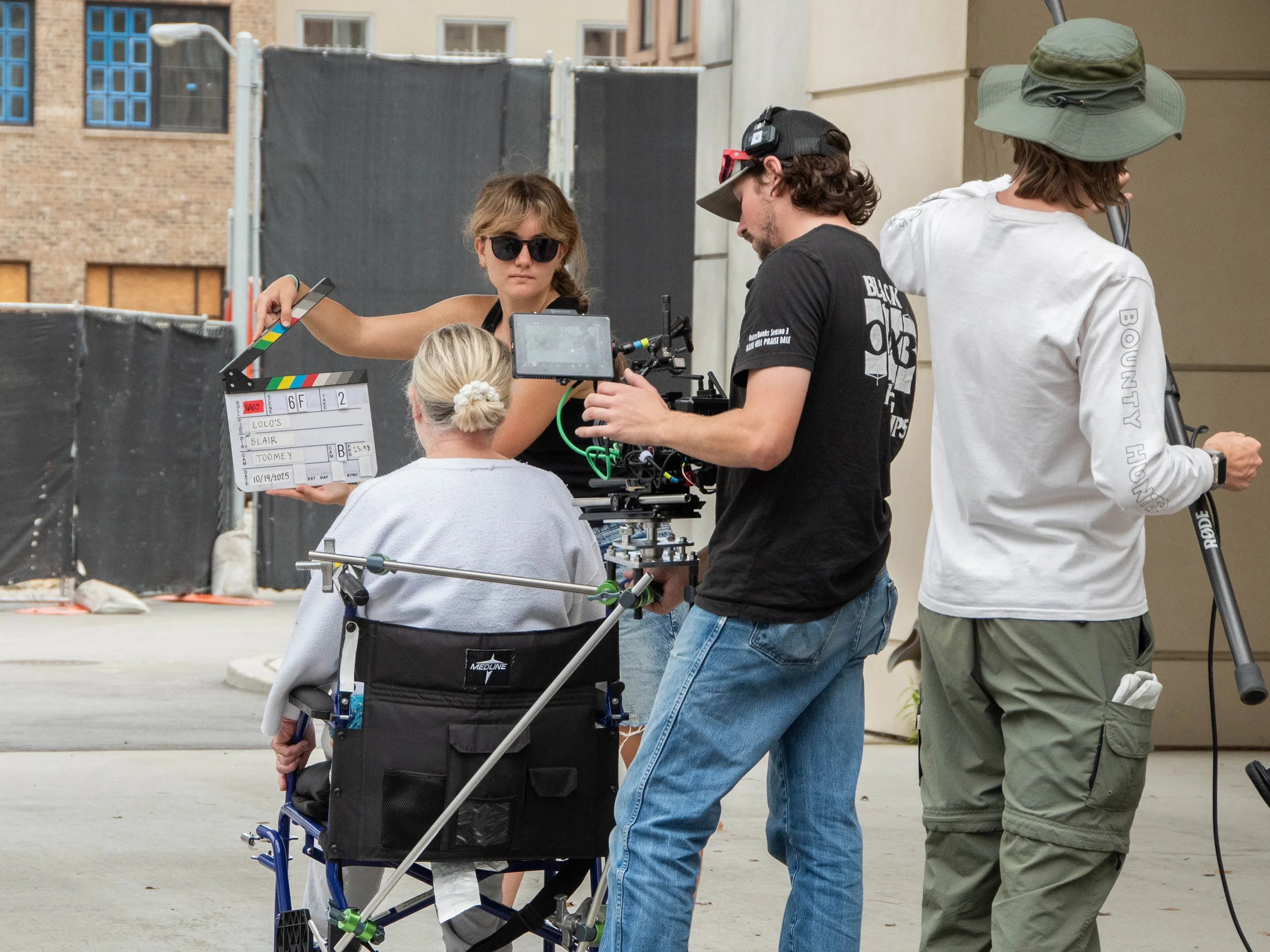 Film crew setting up shooting scene outside, woman in wheelchair holding clapboard, man with camera, another man with equipment, woman with glasses and sunglasses overseeing the scene.