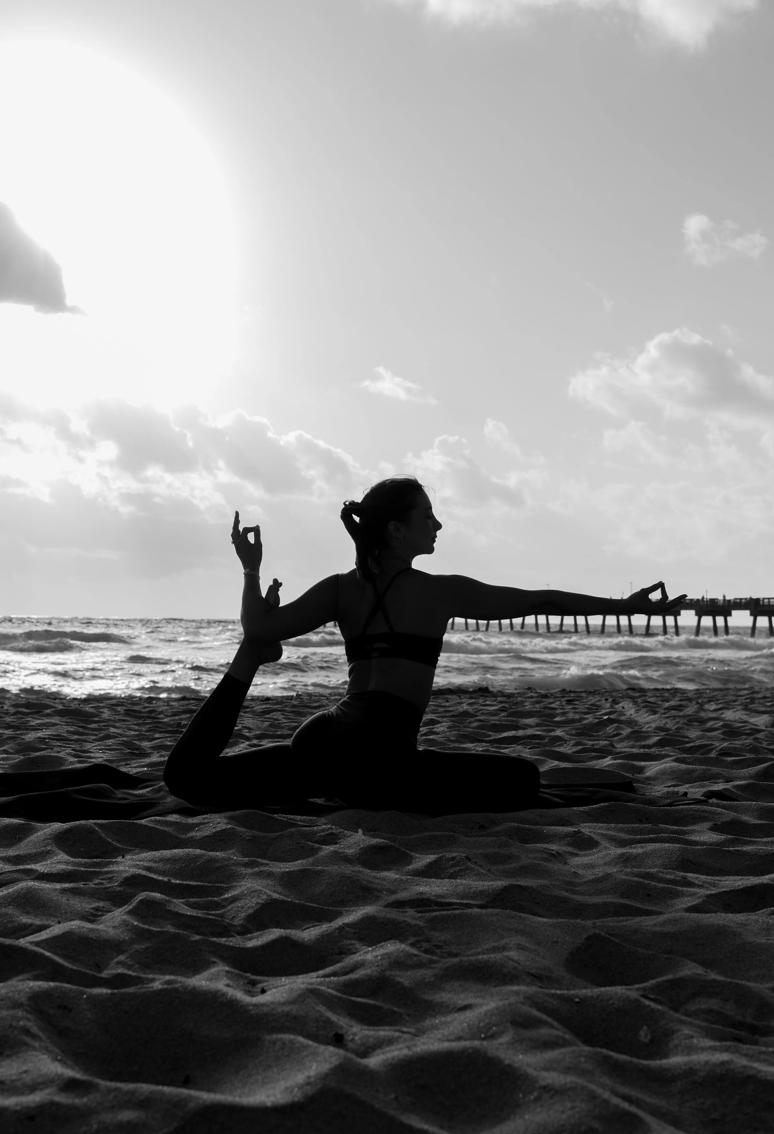 A woman practicing yoga on the beach during sunset or sunrise, silhouetted against the sky with clouds, near a pier.