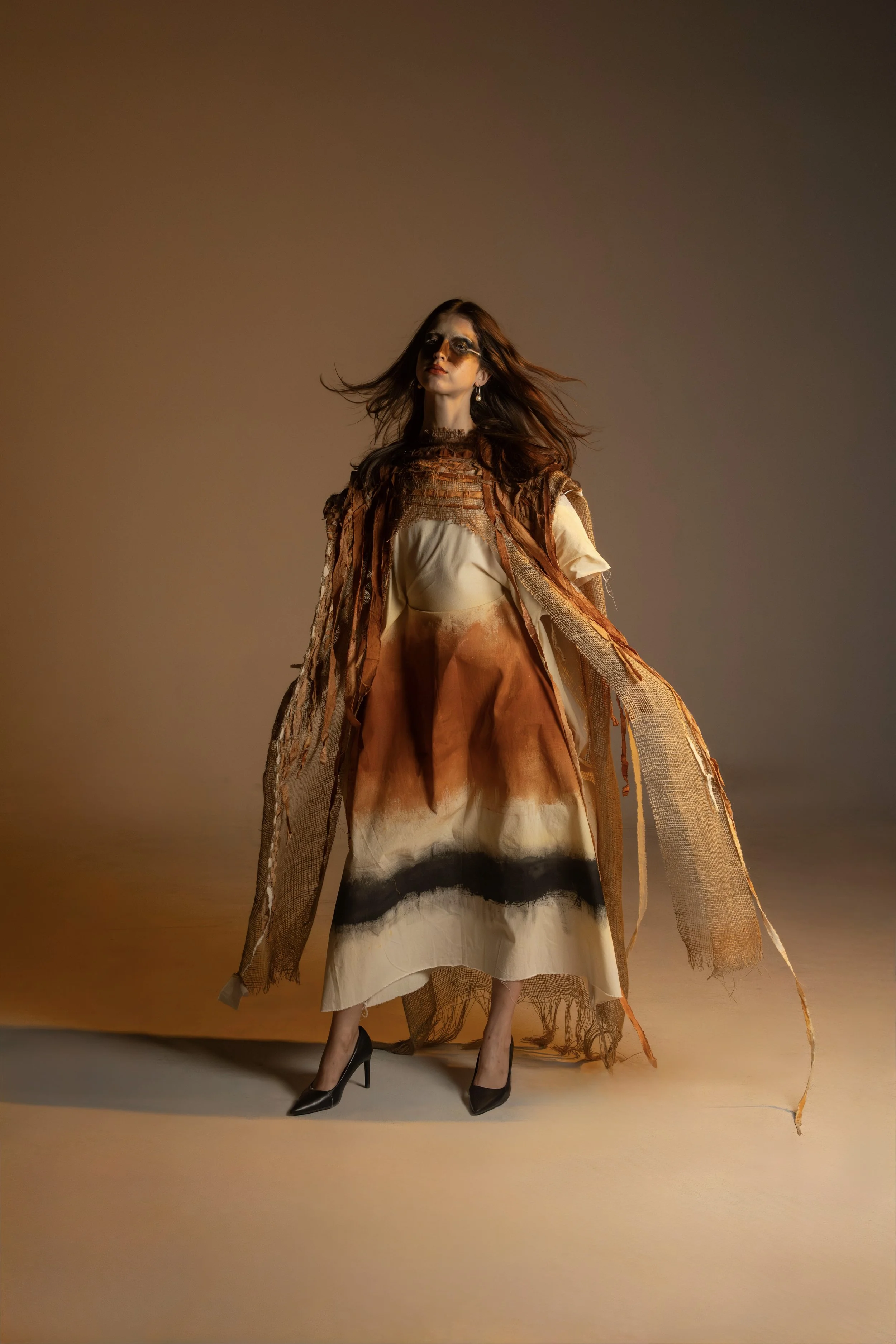 Model with flowing hair in earthy-toned fashion against neutral background, wearing a layered, artistic outfit with a skirt, top, and textured outer layer, and black high heels.
