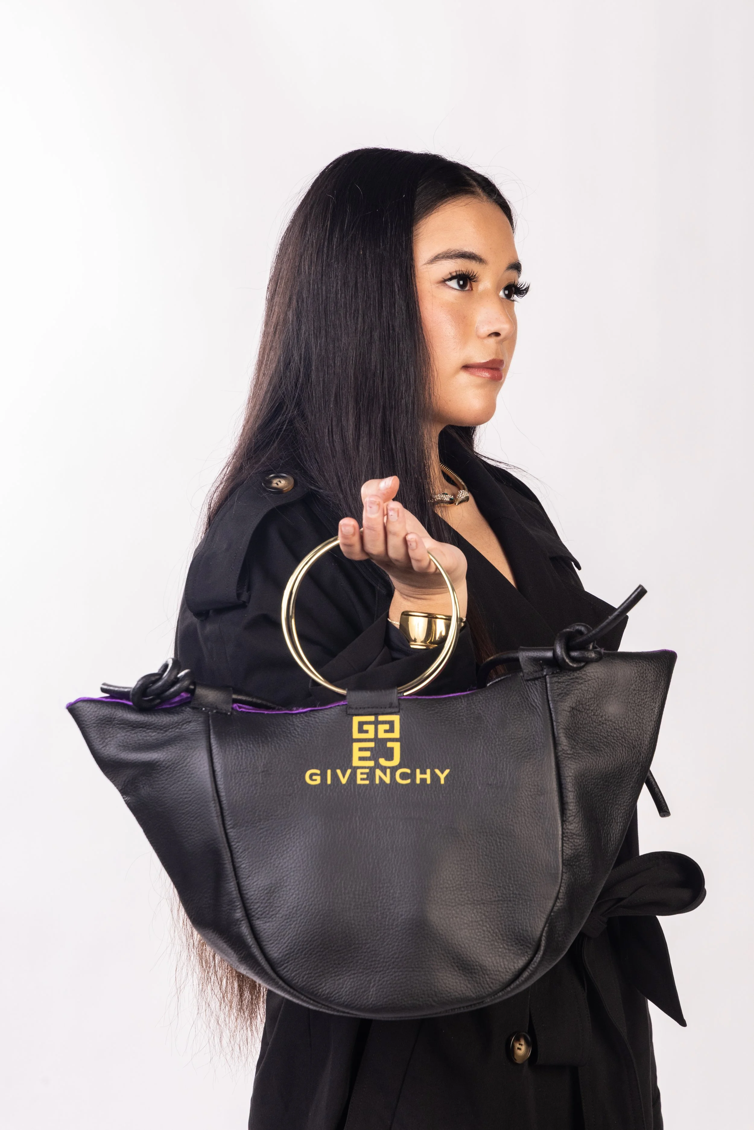 Young woman with long dark hair holding a black Givenchy handbag with gold logo, wearing a black coat and jewelry, against a plain white background.