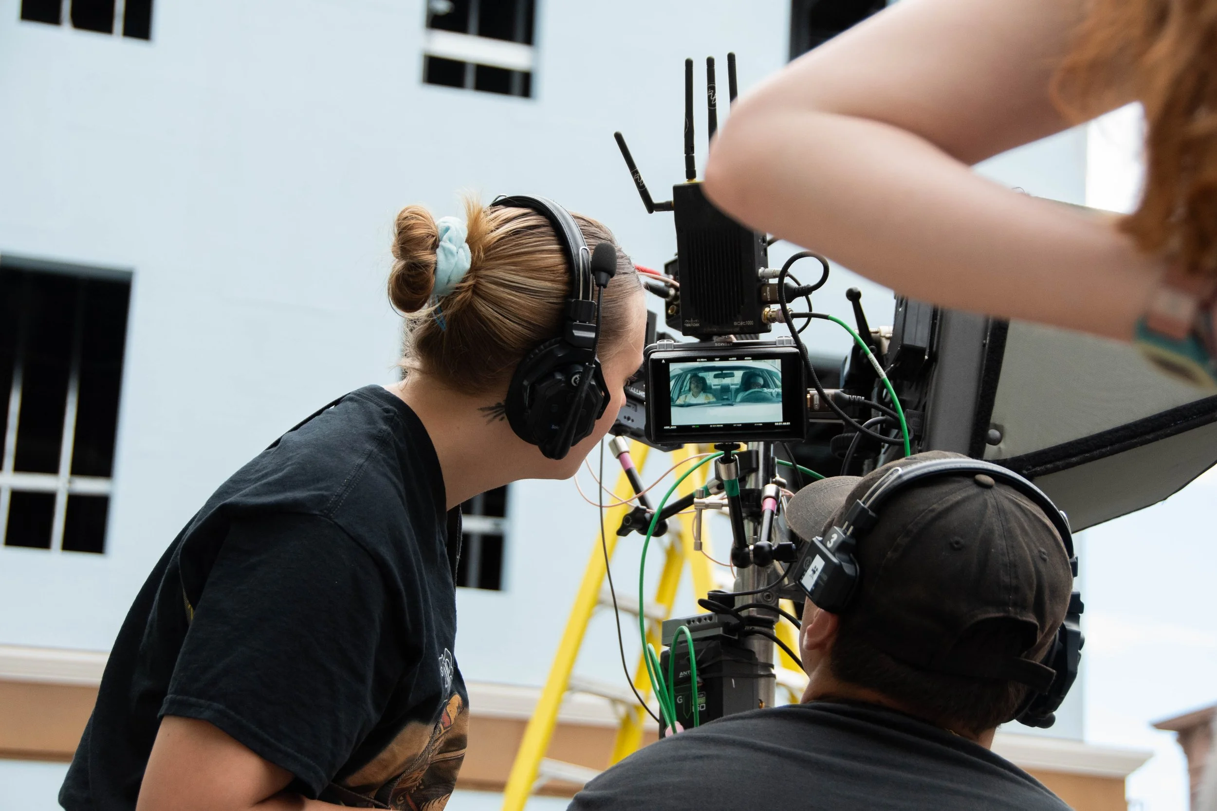 Two crew members working on a film or video camera setup outdoors, with a building and ladders in the background.