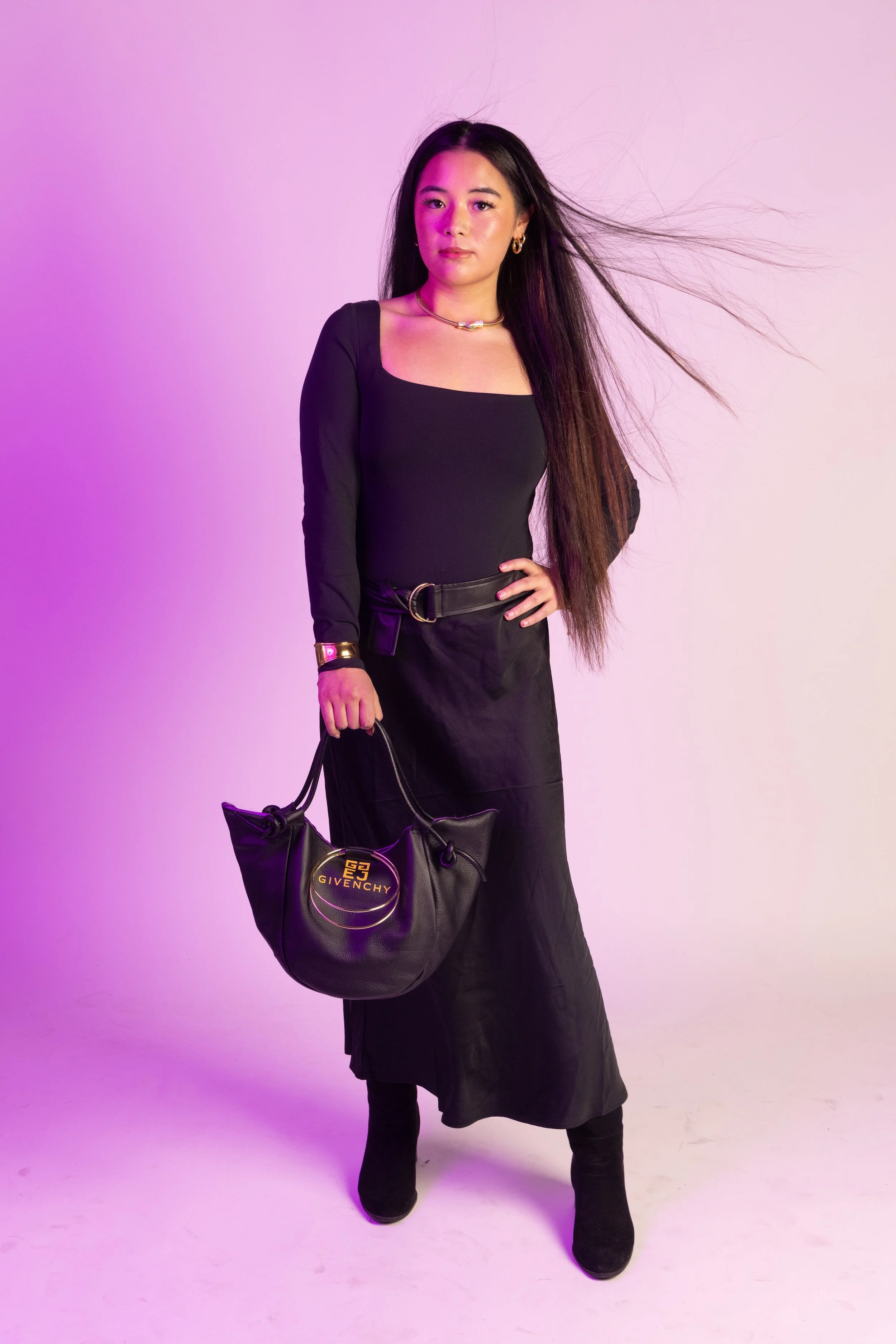 A young woman with long dark hair wearing a black long-sleeve top and a black skirt, holding a black Givenchy handbag, standing against a pink and purple gradient background.