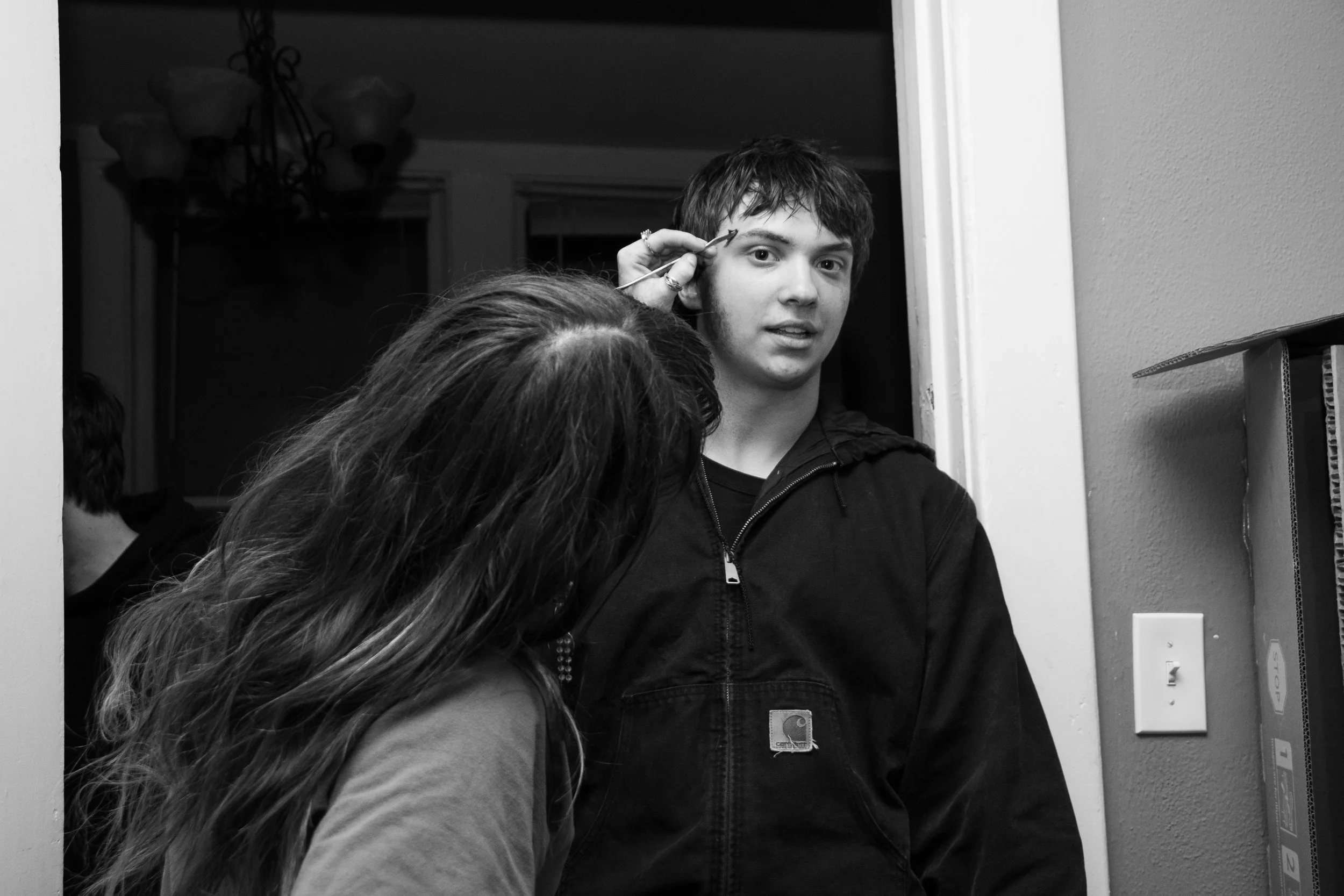 A person is applying makeup to a young man standing in a doorway, with another person partially visible in the background.