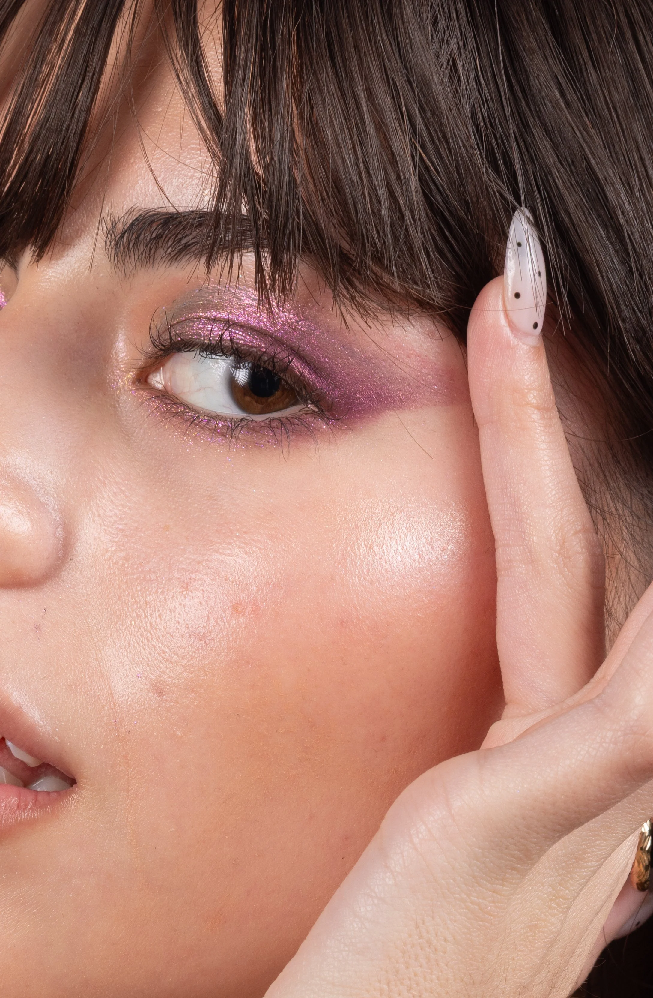 Close-up of a woman's face with pink and purple eyeshadow, long eyelashes, and clear skin. She has brown eyes and is touching her temple with her right hand, which has a white nail with black dots.