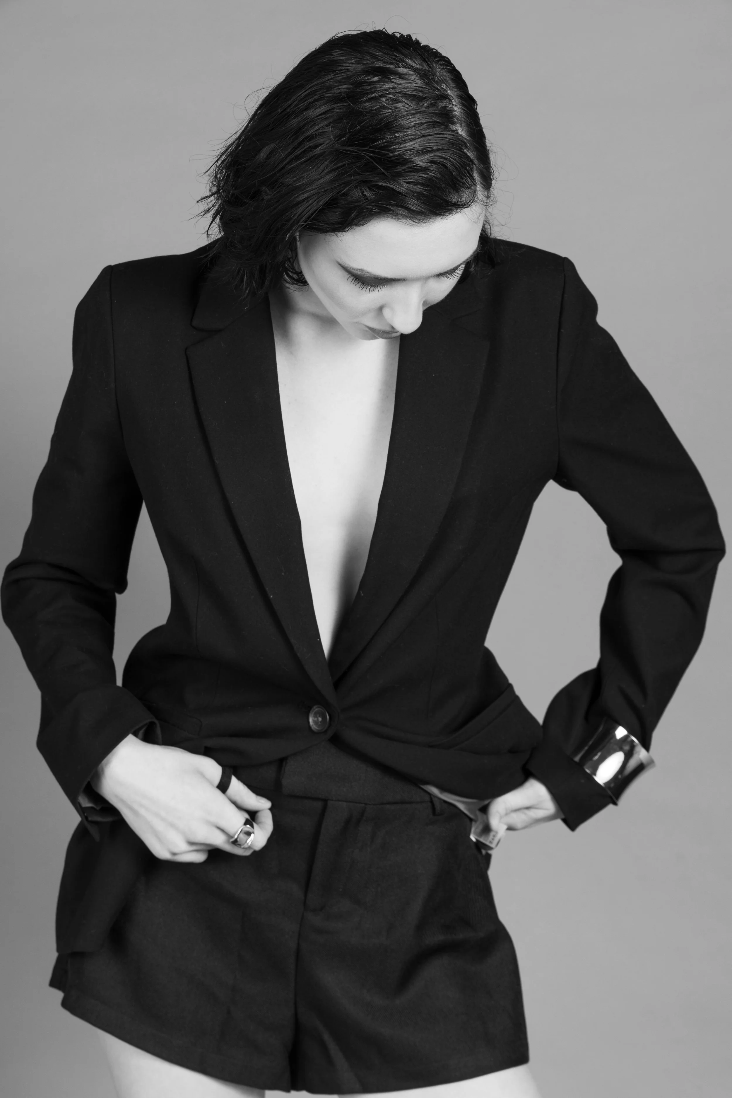 Black and white photo of a woman in a black blazer with rolled-up sleeves, deep V neckline, and matching shorts, posing against a plain gray background. She has dark, wavy, shoulder-length hair and is looking downward with her hands in her pockets.