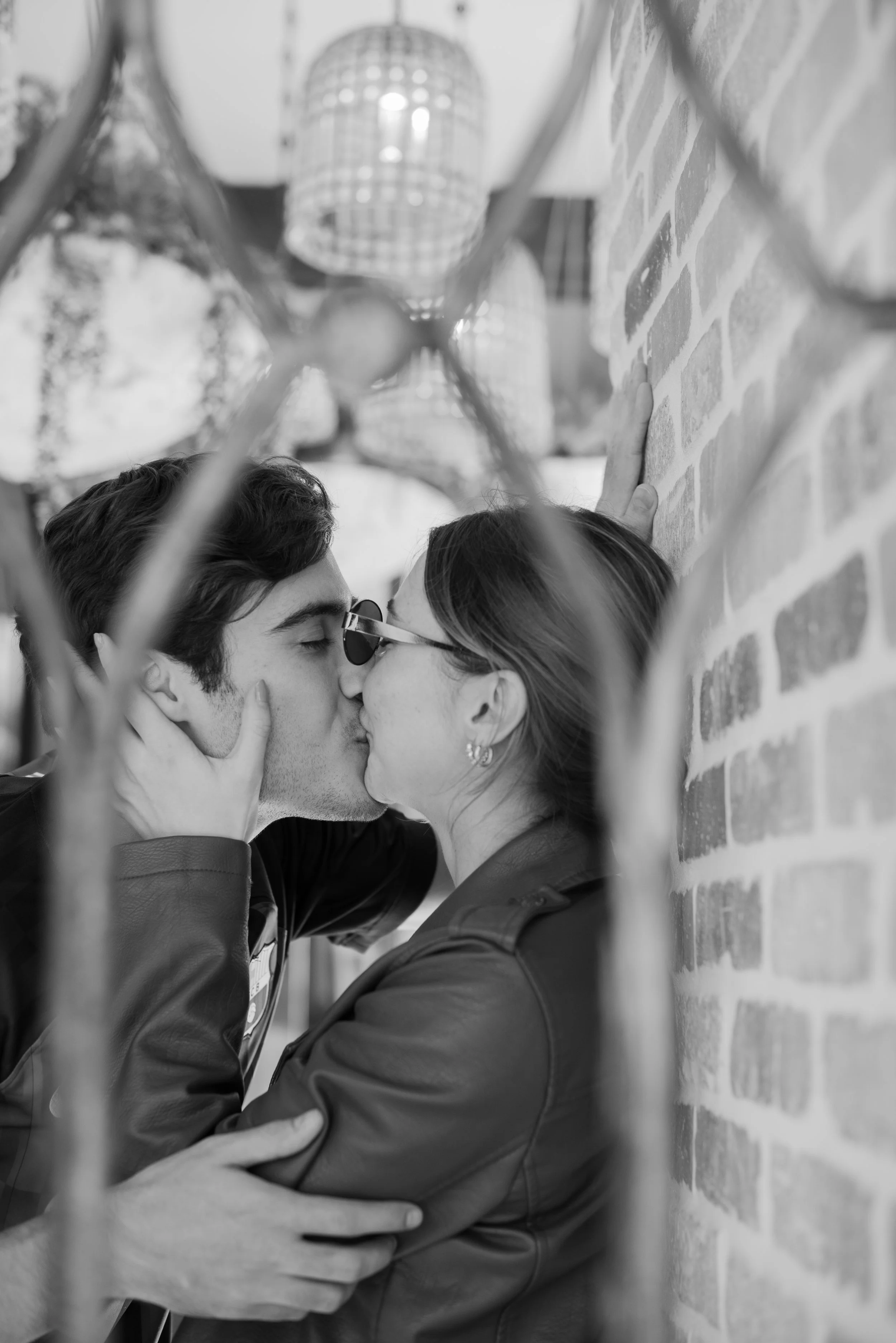 A black and white photo of a couple kissing closely, with their hands touching each other's faces and necks, framed by what appears to be a fence or railing in the foreground. The scene takes place against a brick wall, with hanging light fixtures ab