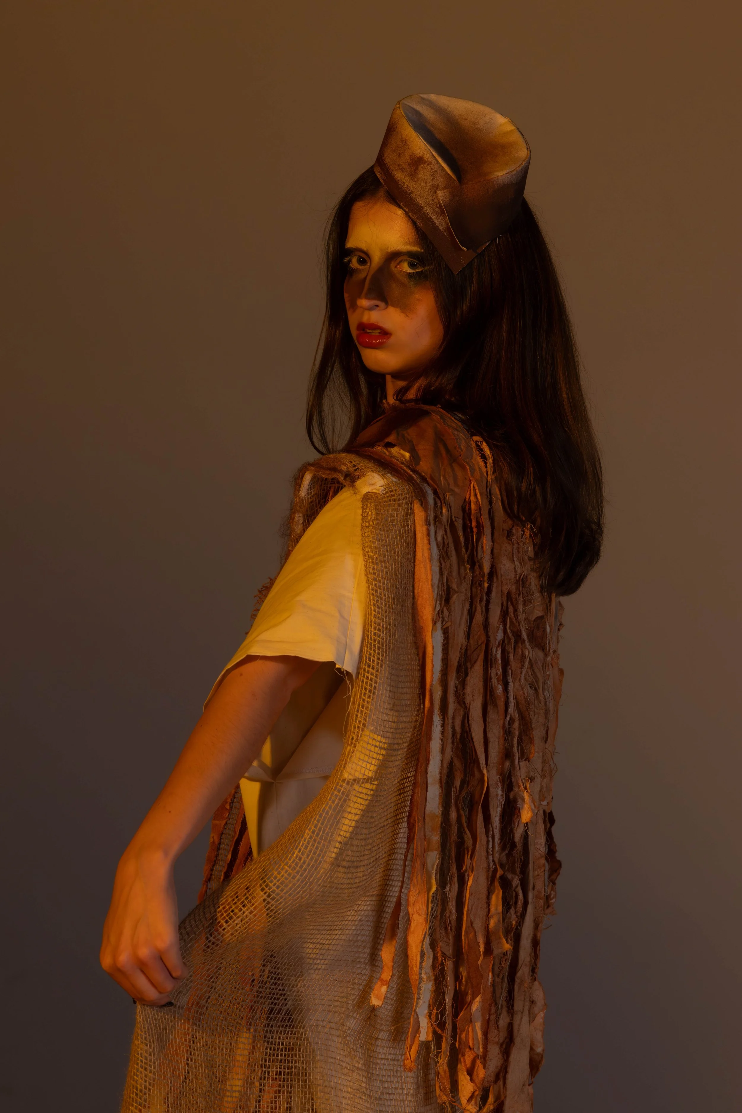 A woman wearing a brown paper hat and a textured, layered brown and beige outfit stands against a plain background, looking over her shoulder.