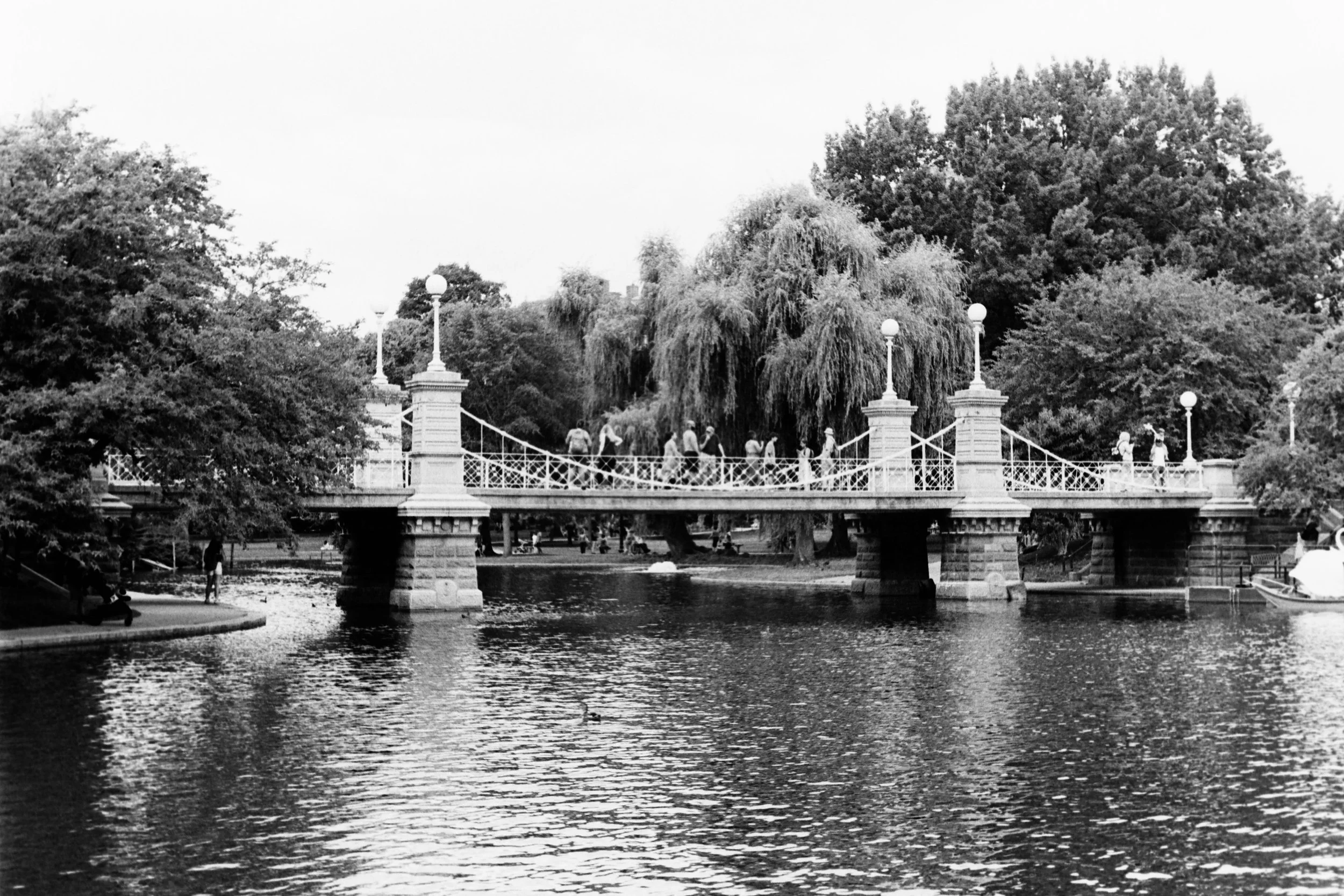 Black and white photo of a small park with a bridge over a pond, surrounded by trees and people walking across the bridge.