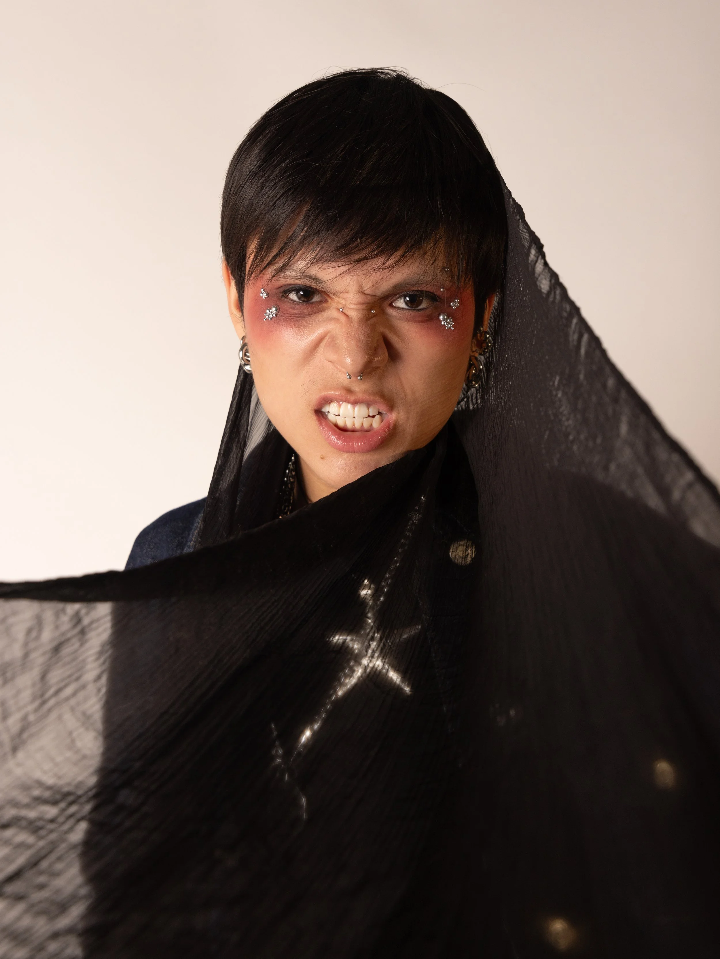 Person with short dark hair, facial piercings, and facial jewelry, making an aggressive facial expression, partially covered in black sheer fabric, against a plain background.