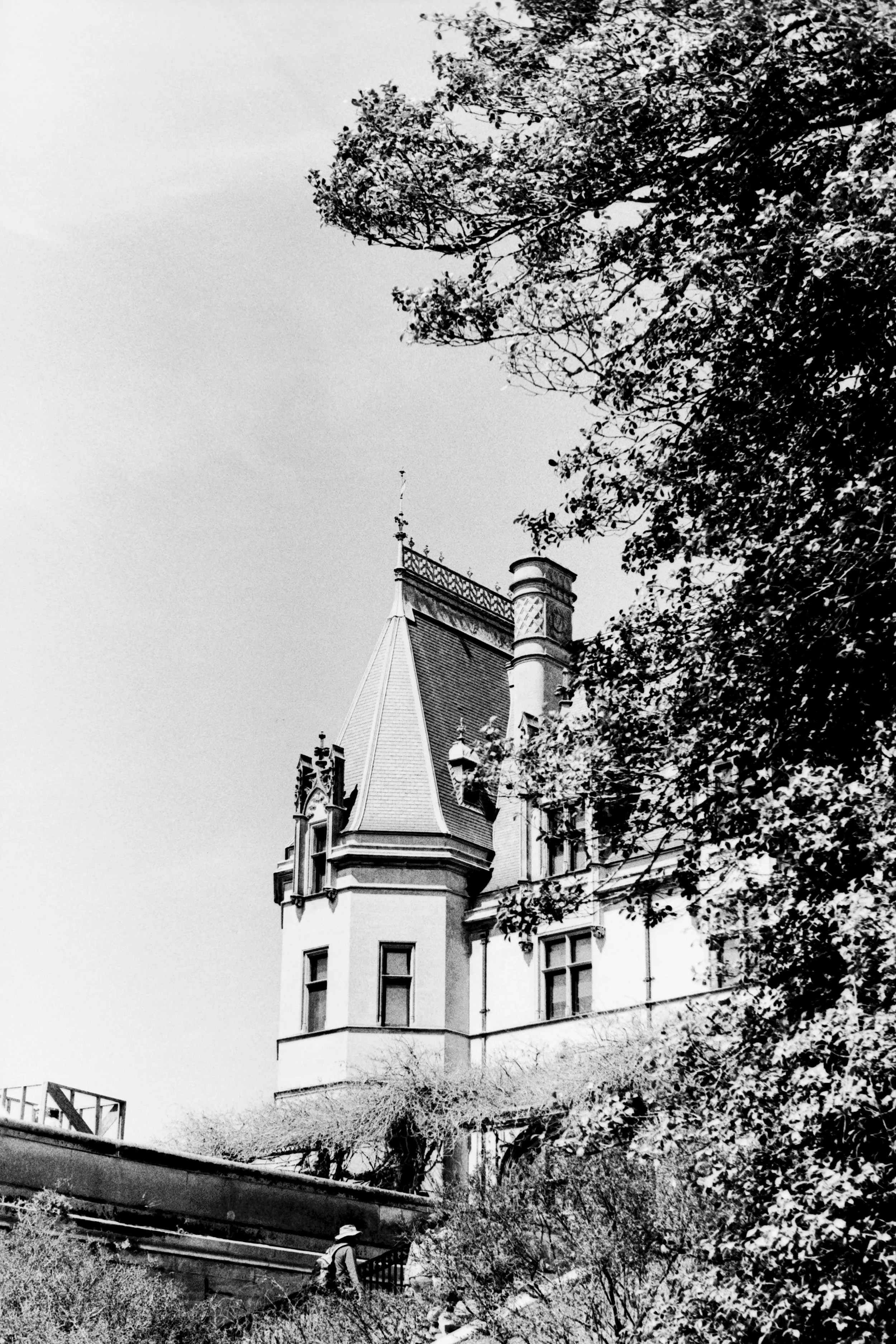 A black and white photograph of a castle-like building with a steep roof, turrets, and ornate architectural details, partially obscured by trees.