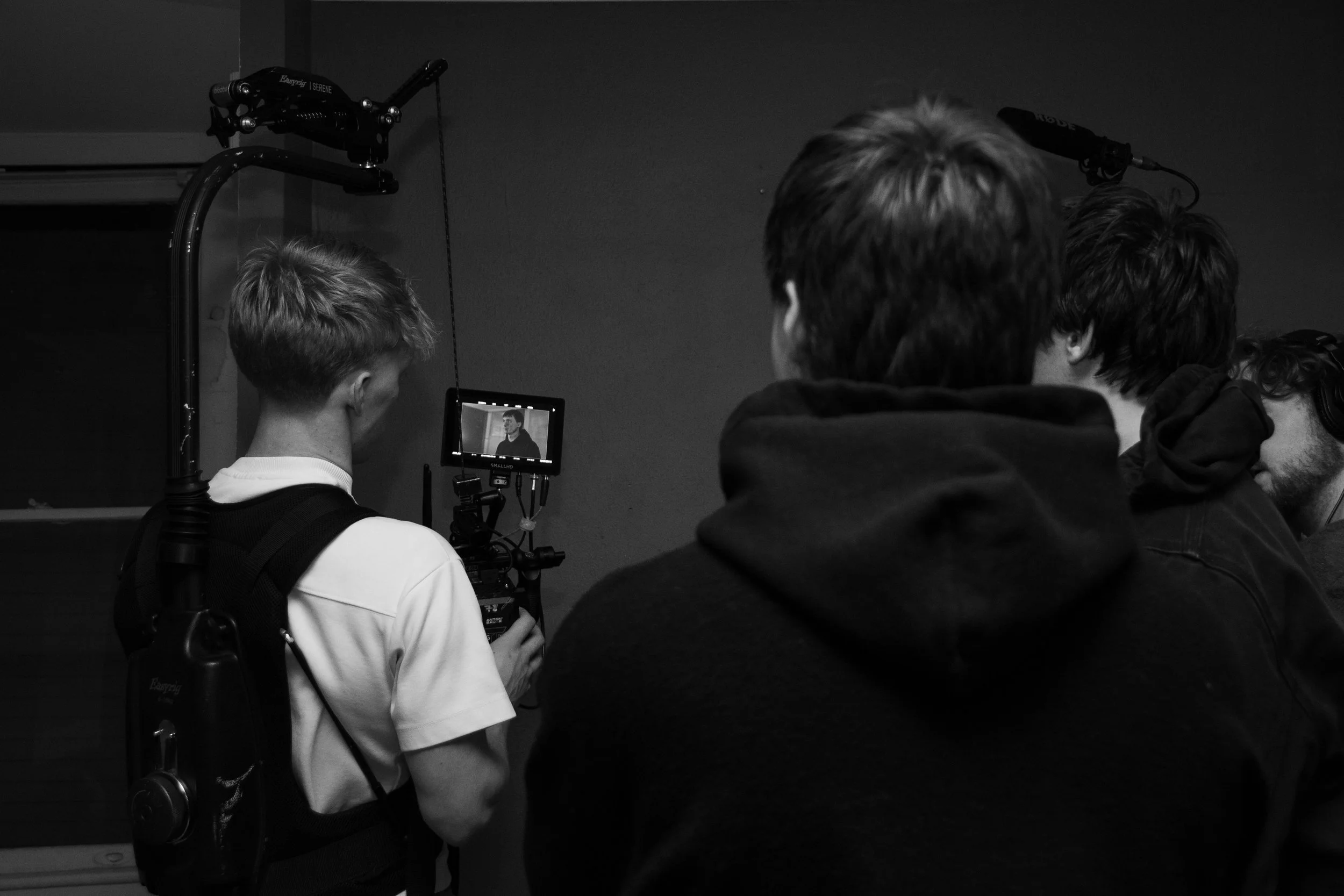 A person operating a film camera with a monitor, being watched by a group of individuals, in a dimly lit room, black and white photo.