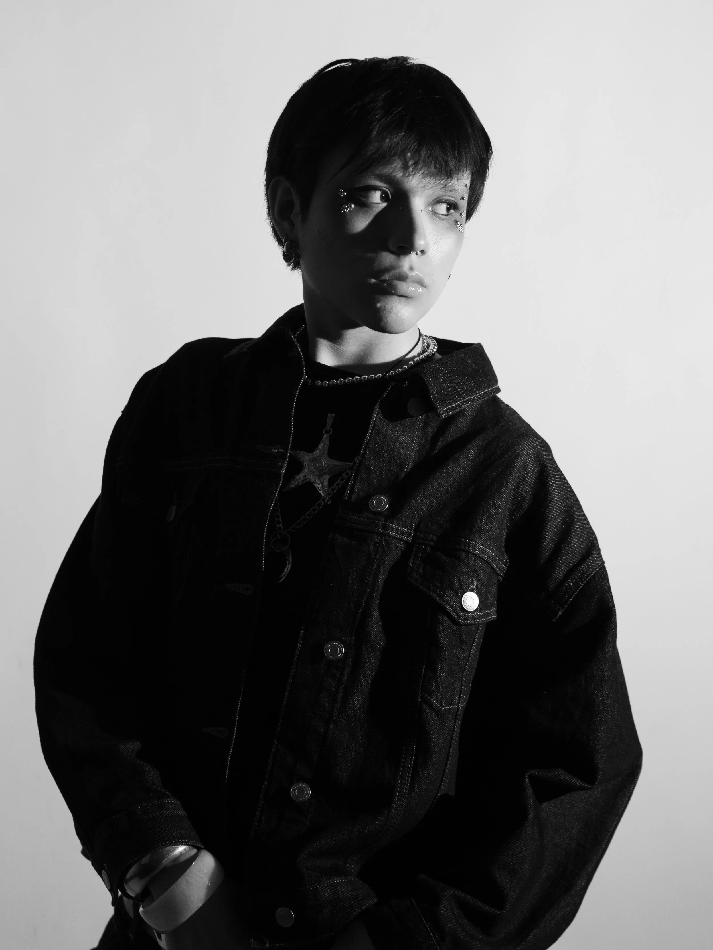 Black and white portrait of a young woman with short hair, wearing a denim jacket and jewelry, looking to the side against a plain background.