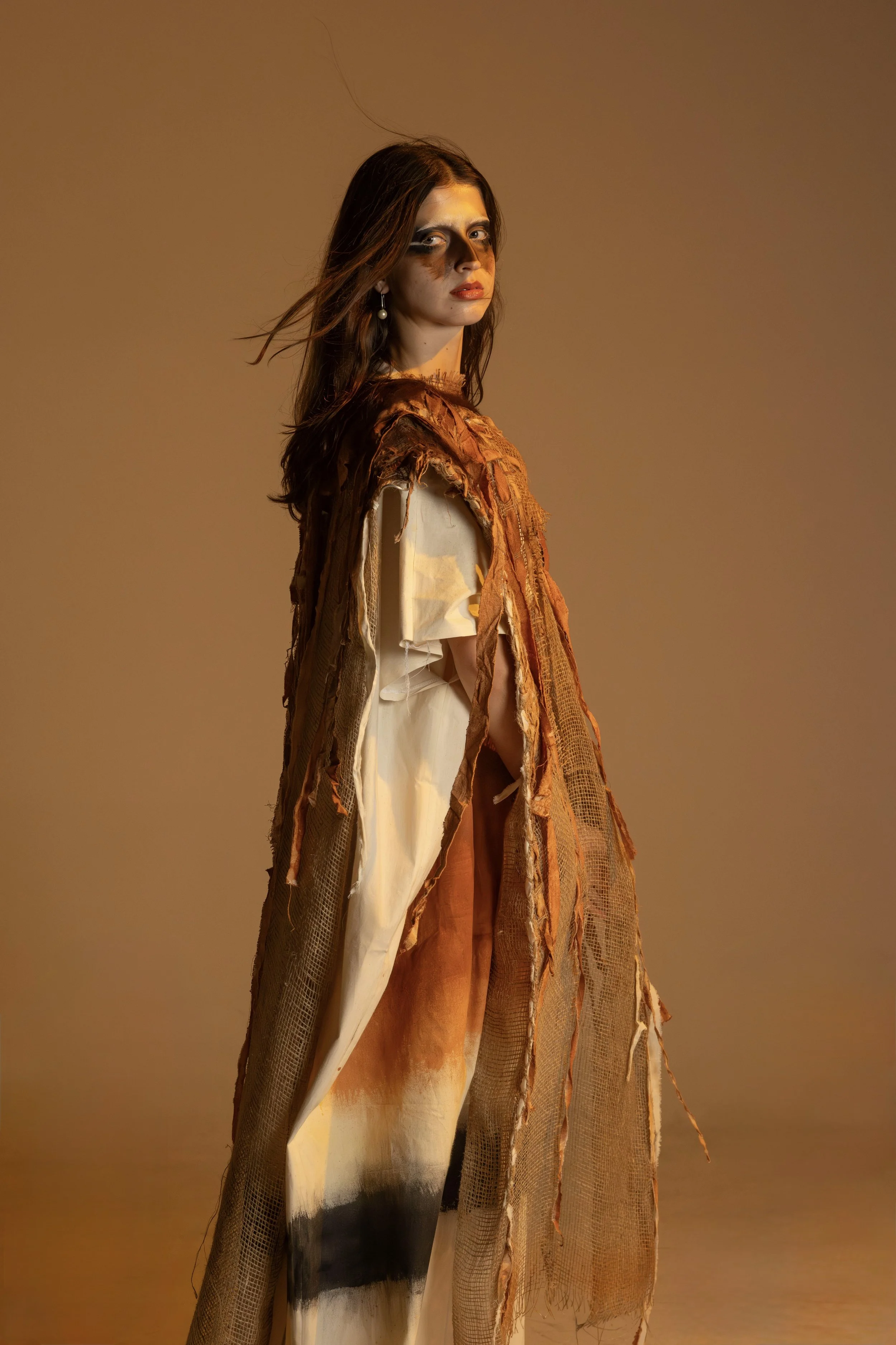 A woman with dark hair and makeup in brown and beige tones, wearing an avant-garde, textured orange and beige layered outfit, posed against a warm, plain background.