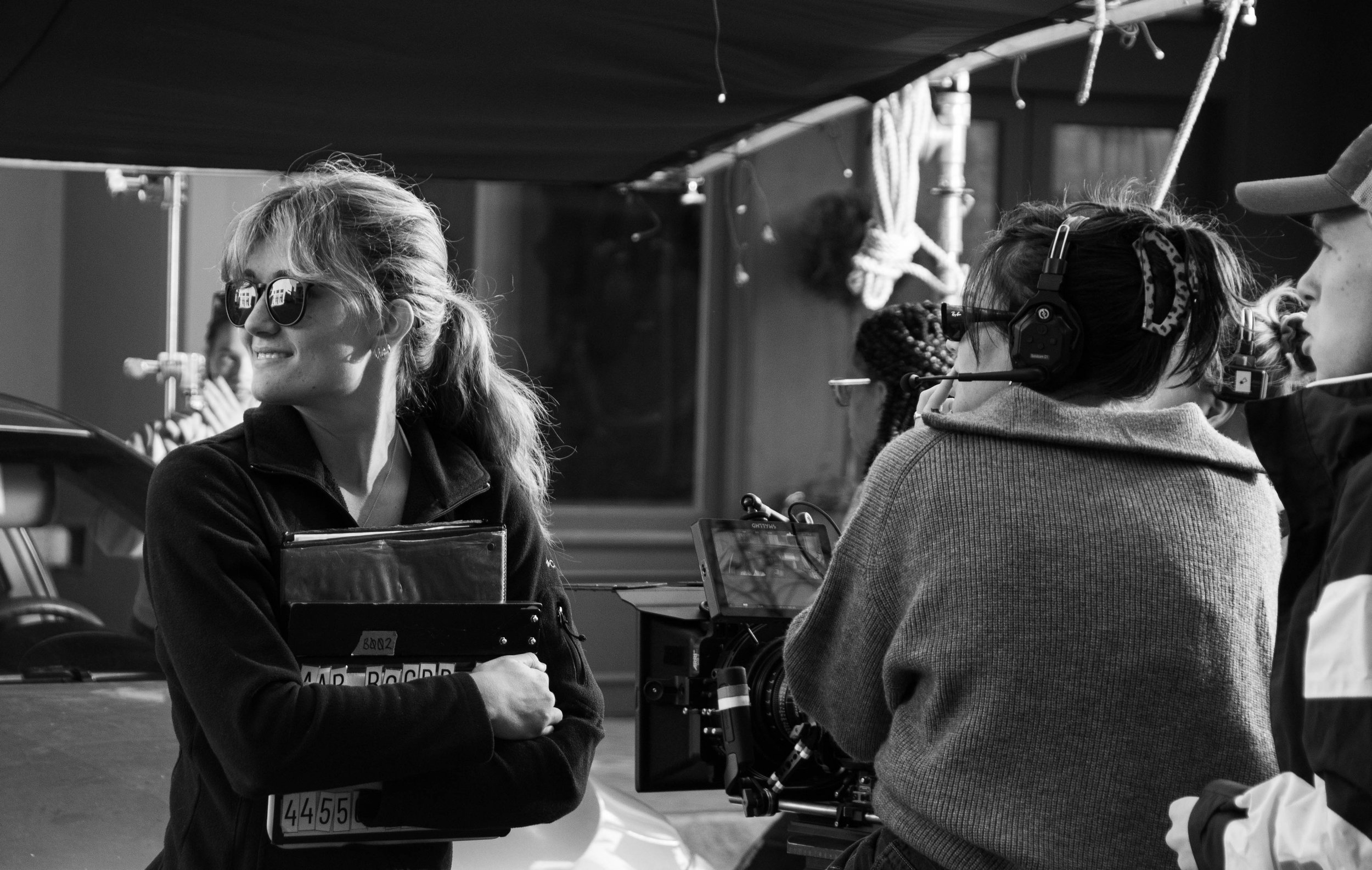 A group of people on a film or television set. A woman with glasses and a smile is holding a clipboard, while a woman with headphones operates a camera. Others are standing nearby, engaged in the production.
