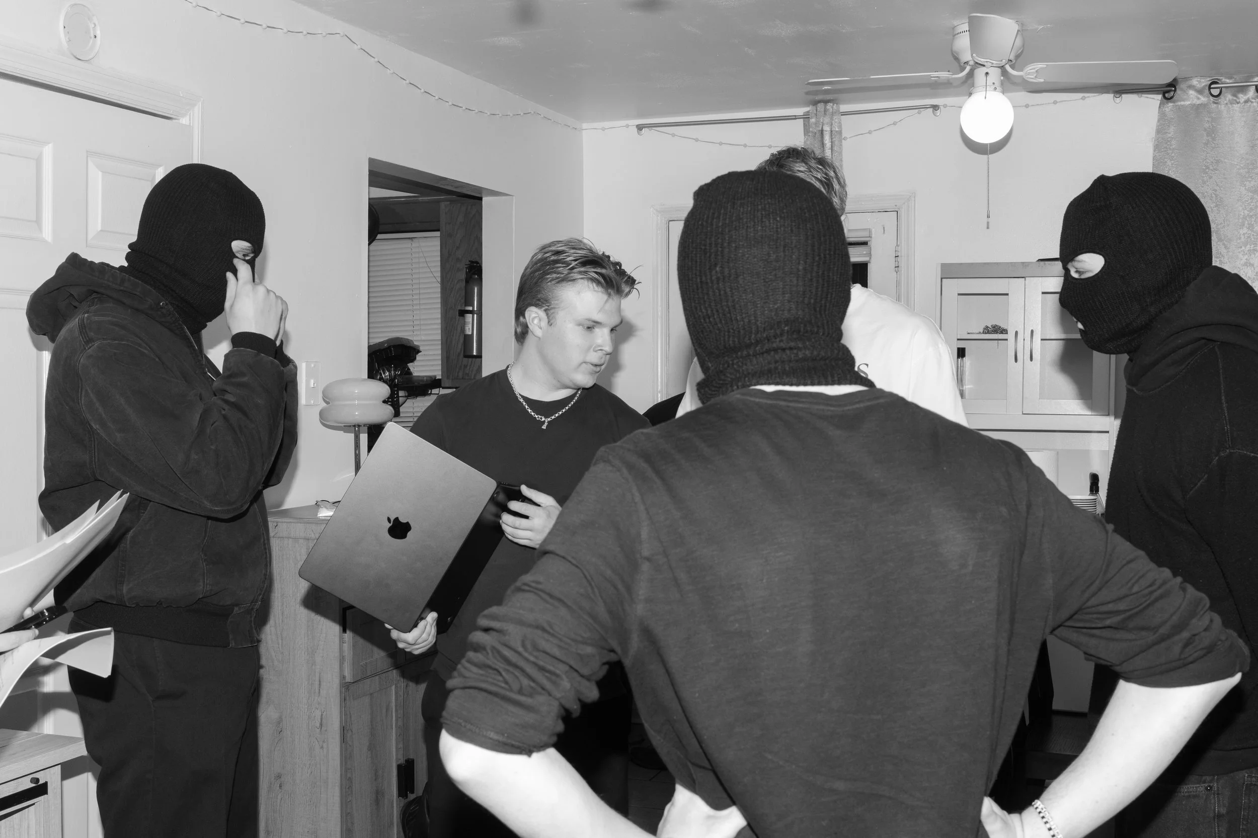 A group of five young men in a kitchen, some wearing ski masks, gathered around a person holding a laptop. The kitchen has cabinets, a ceiling fan, and string lights.