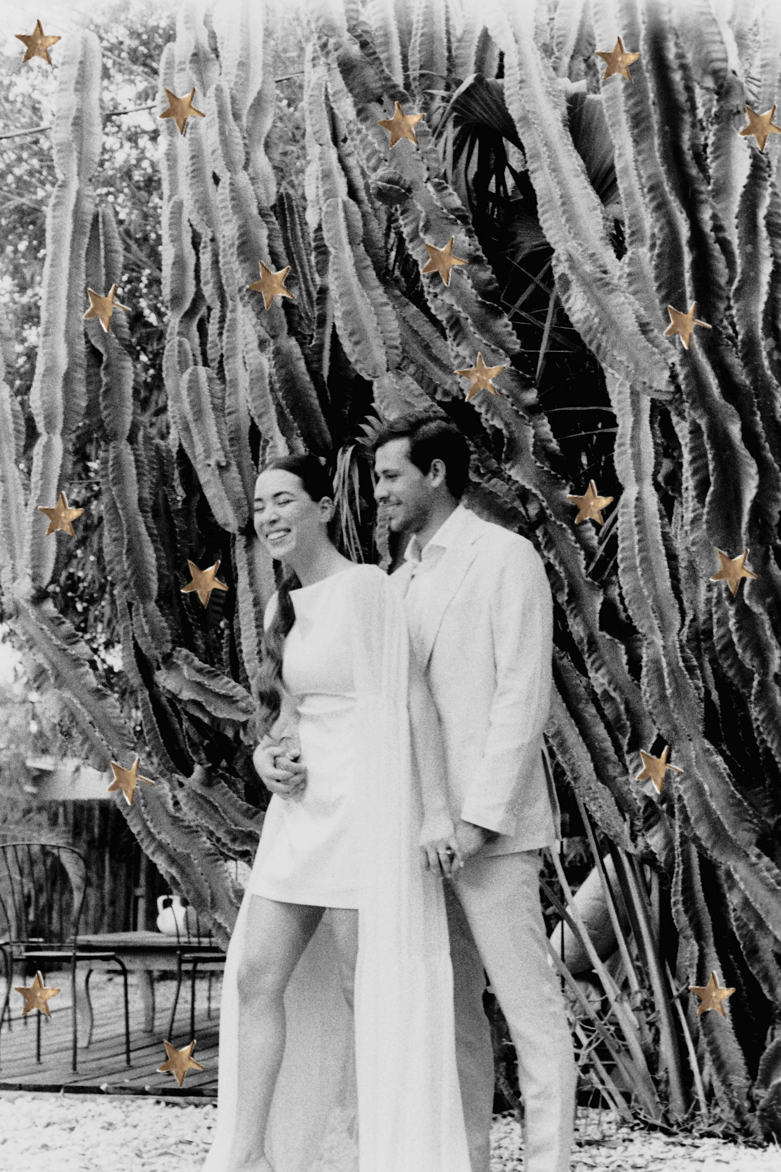 A black-and-white photograph features a smiling woman in a short, white dress and a man in a light-colored suit standing outside in front of a large cactus plant. The woman has long dark hair and is holding her partner's hand, while the man has dark 