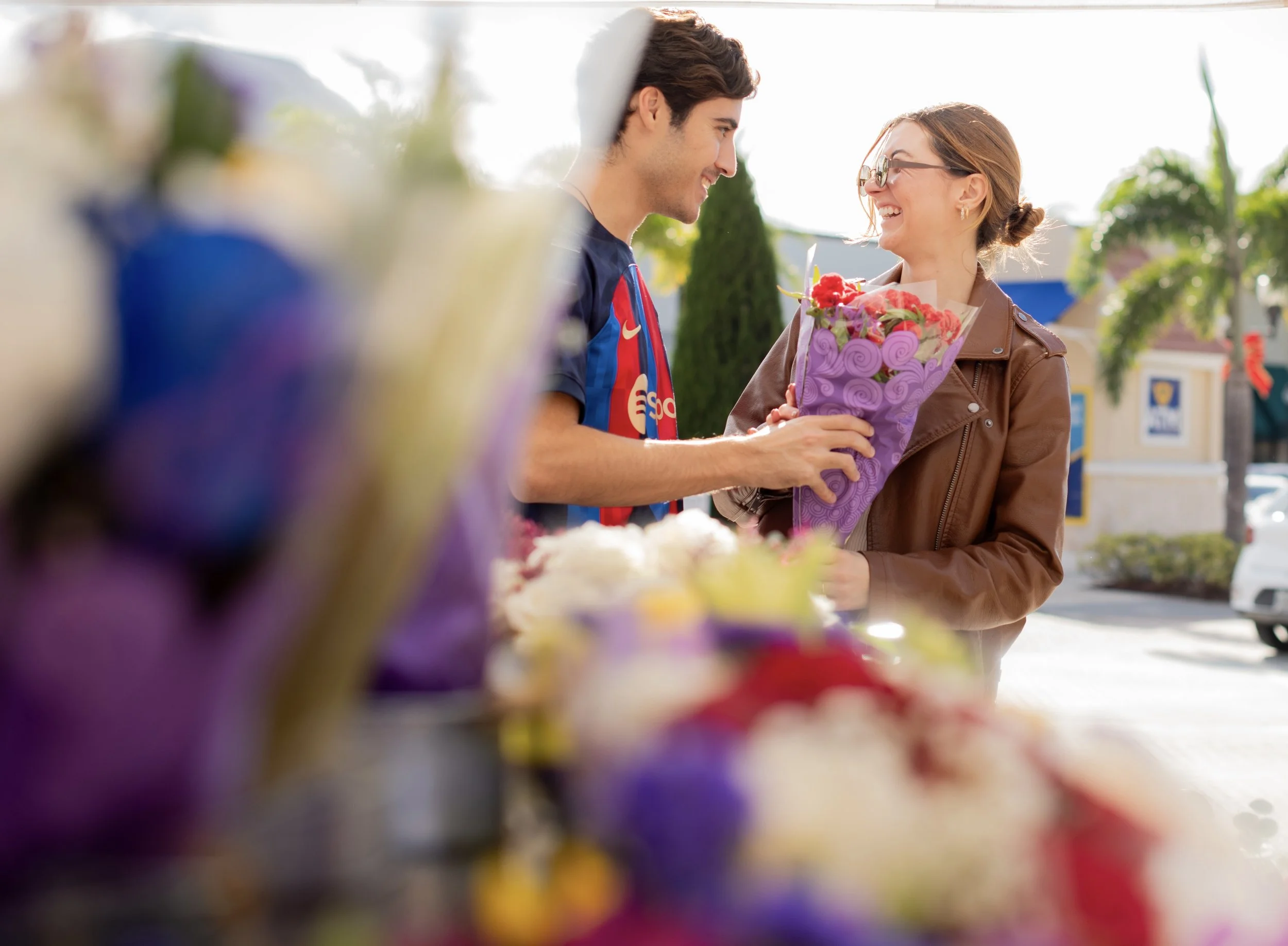 A young man giving a bouquet of flowers to a young woman who is smiling. They are outdoors with a blurred background of a small shopping area and greenery.
