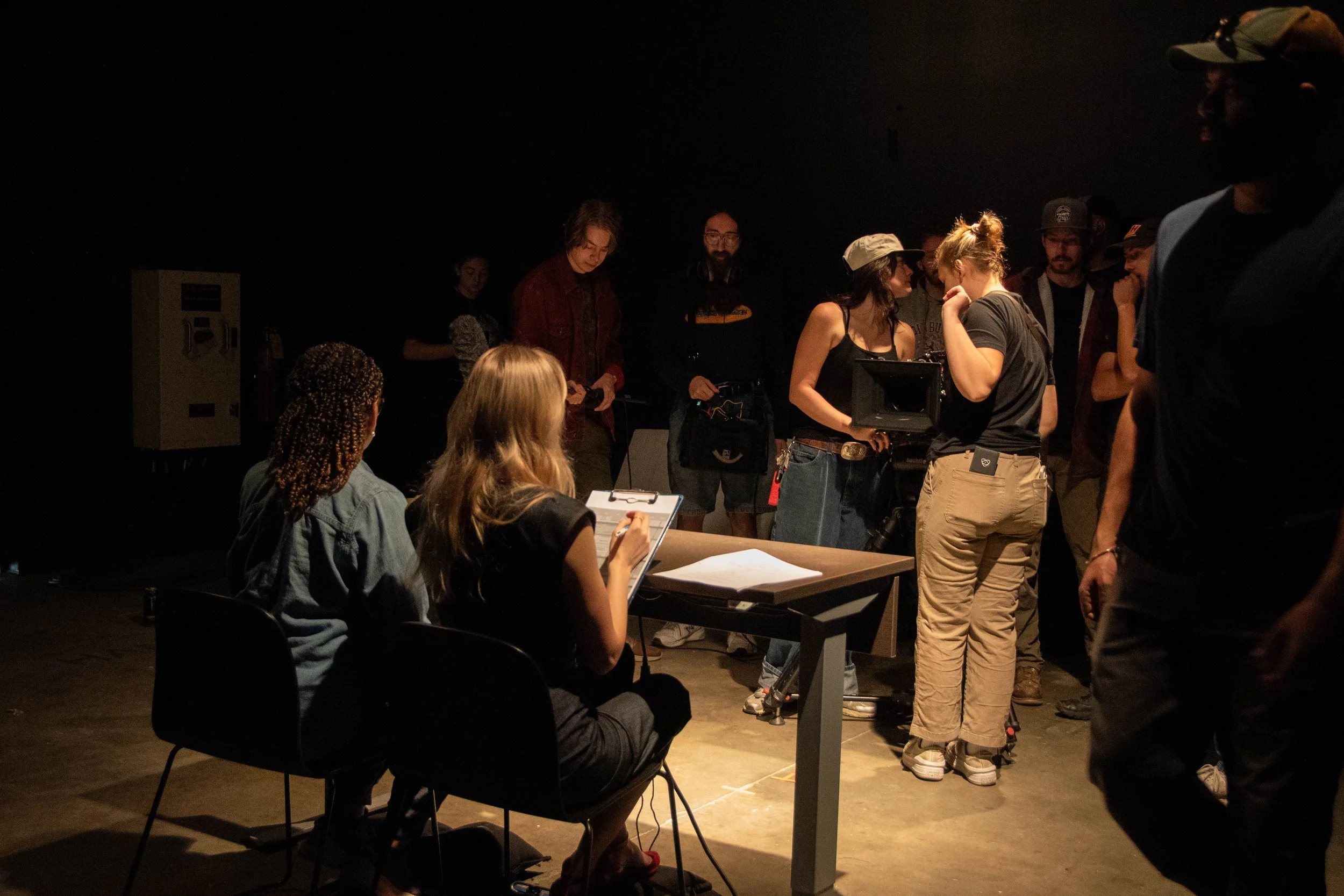 Filmmaking crew working on set in a dark room, with two women sitting at a table in the foreground and multiple people gathered around a camera and equipment in the background.