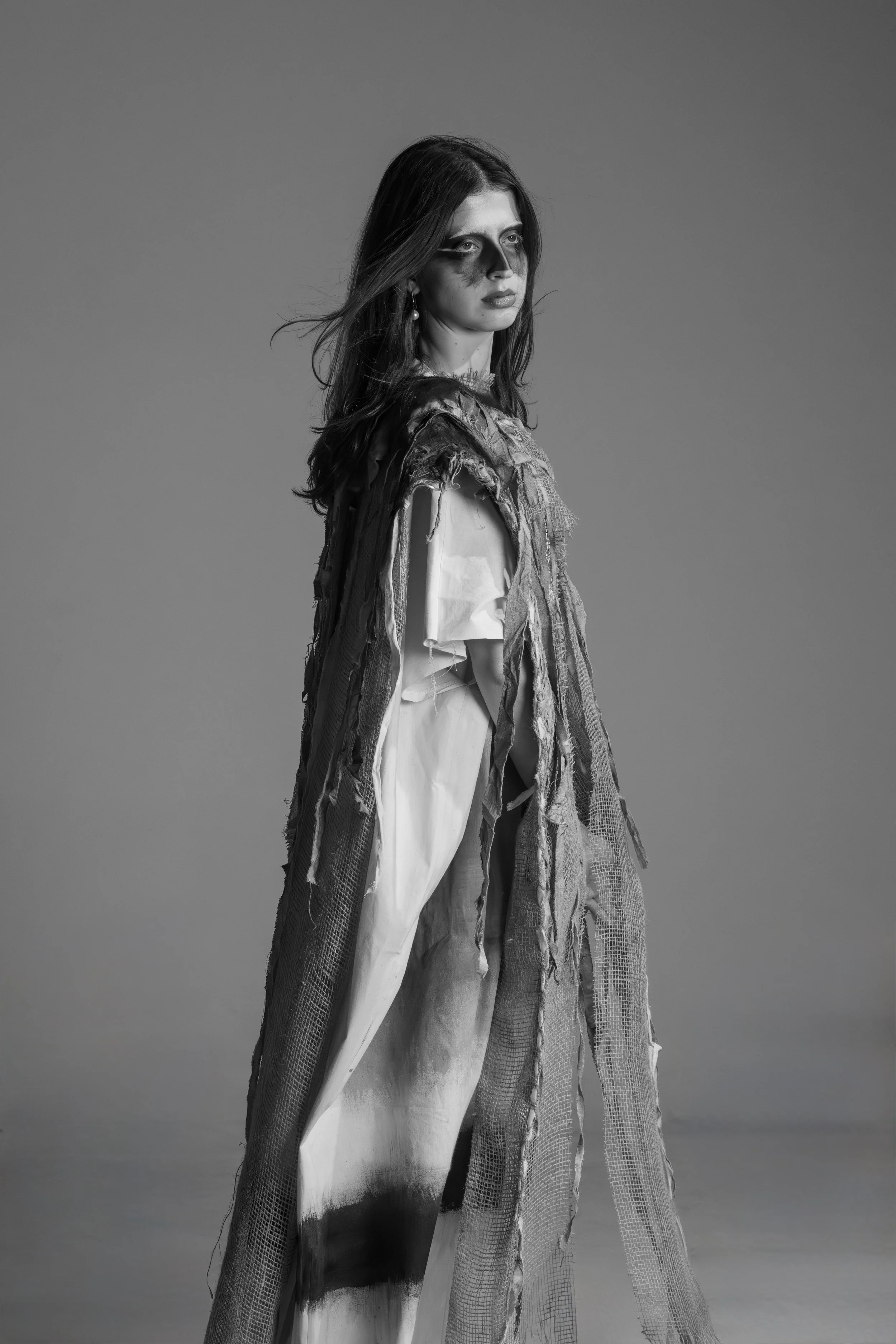 Black and white photo of a woman with long hair, wearing a distressed and layered outfit, standing against a plain backdrop.