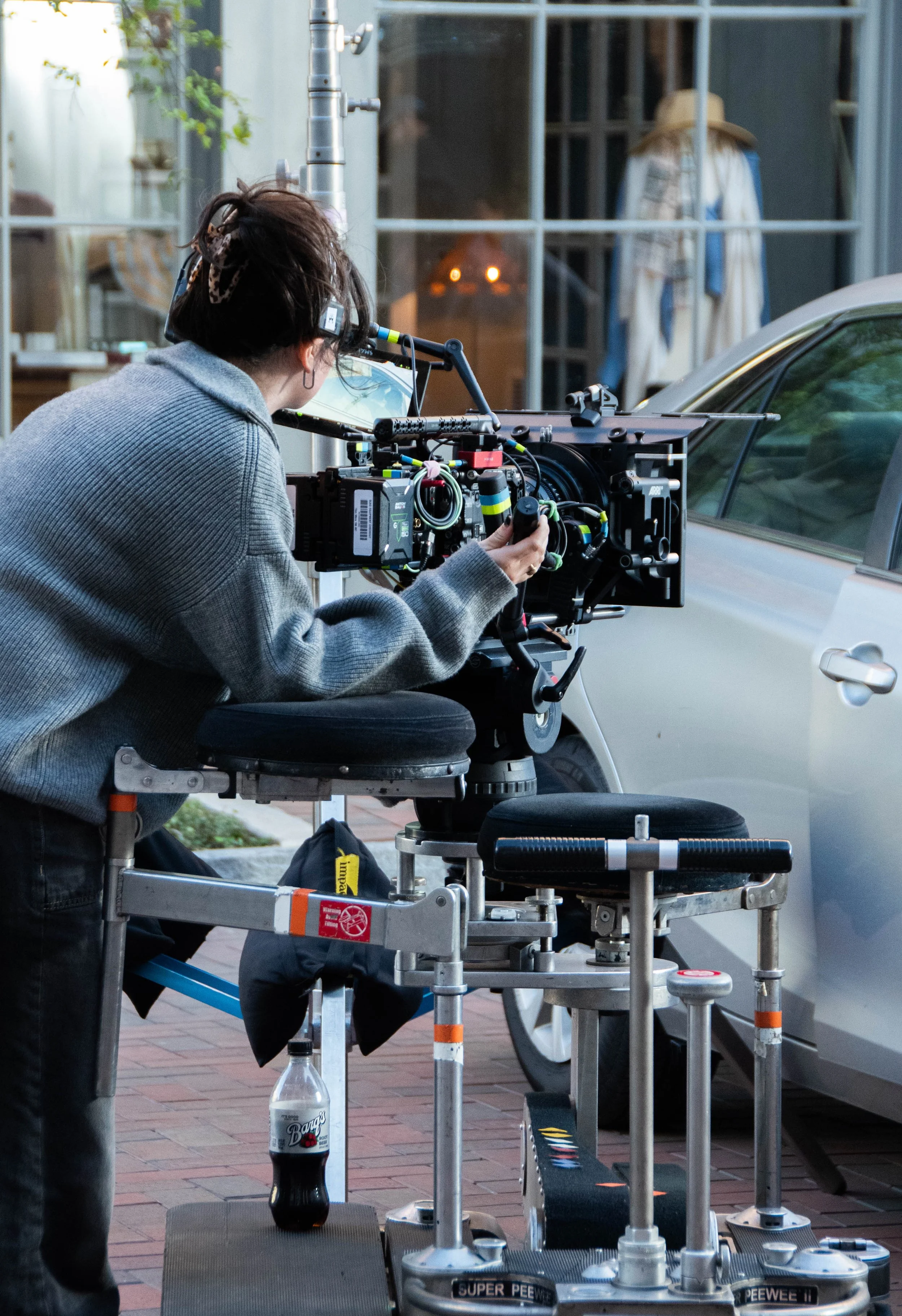A film crew member operating a camera on a dolly during an outdoor shoot on a sidewalk, with a parked car and a woman in a wide-brimmed hat in the background.